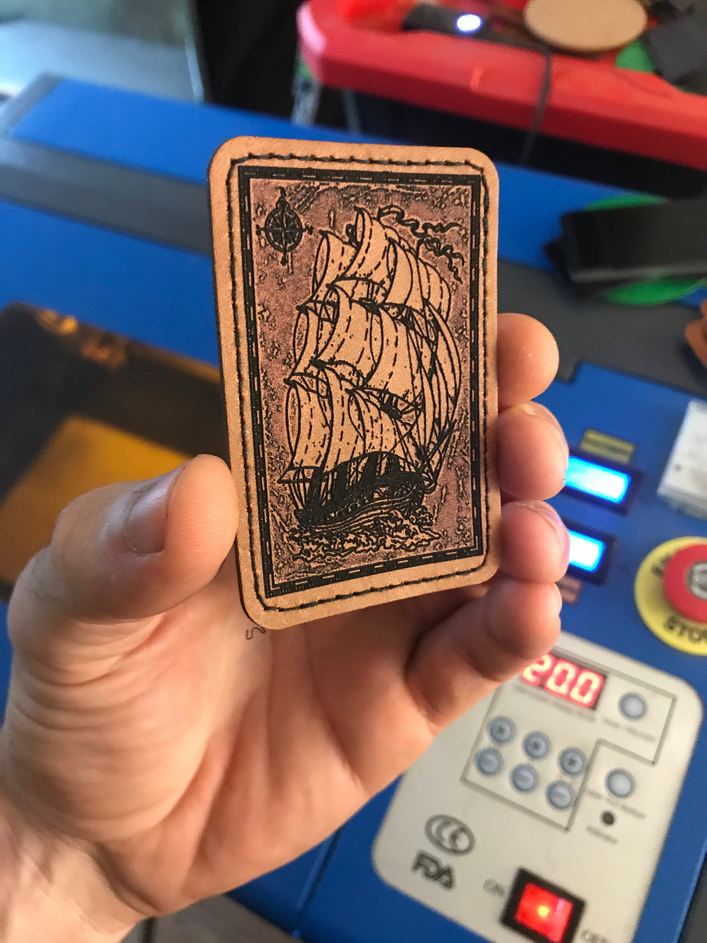 Hand holding a laser engraved leather patch with a sailship design against a blurred background