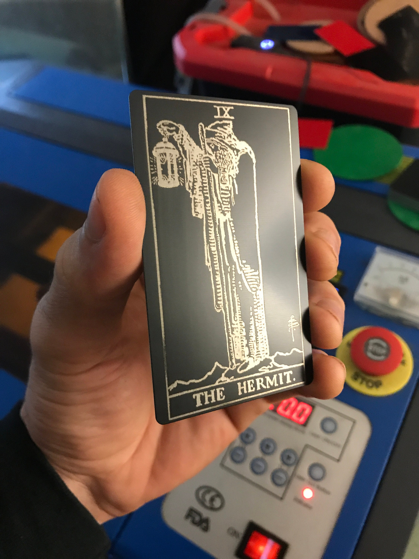 Hand holding a silver/black metal tarot card titled 'The Hermit' with a colorful background.