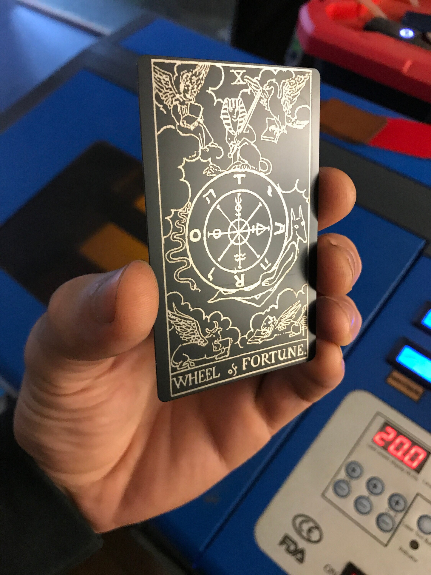 Hand holding an anodized aluminum matte black/silver laser engraved keepsake card with 'Wheel of Fortune' design against a blurred background