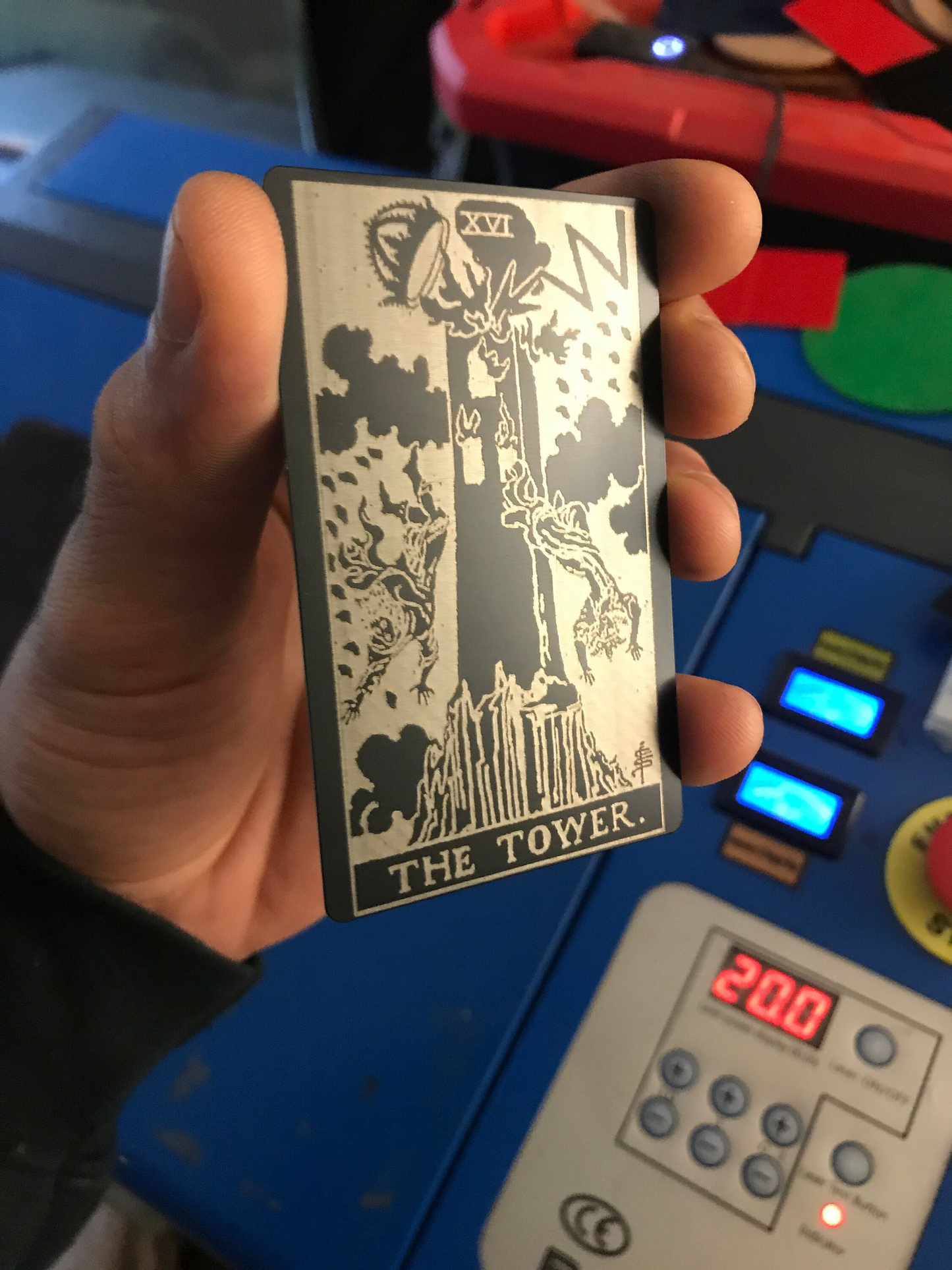 Hand holding a metal laser engraved tarot card titled 'The Tower' in front of a control panel.