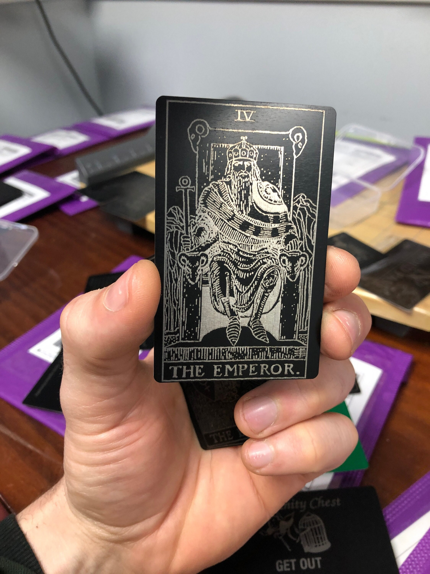 Hand holding a metal black/silver tarot card titled 'The Emperor' with a blurred background of more tarot cards.