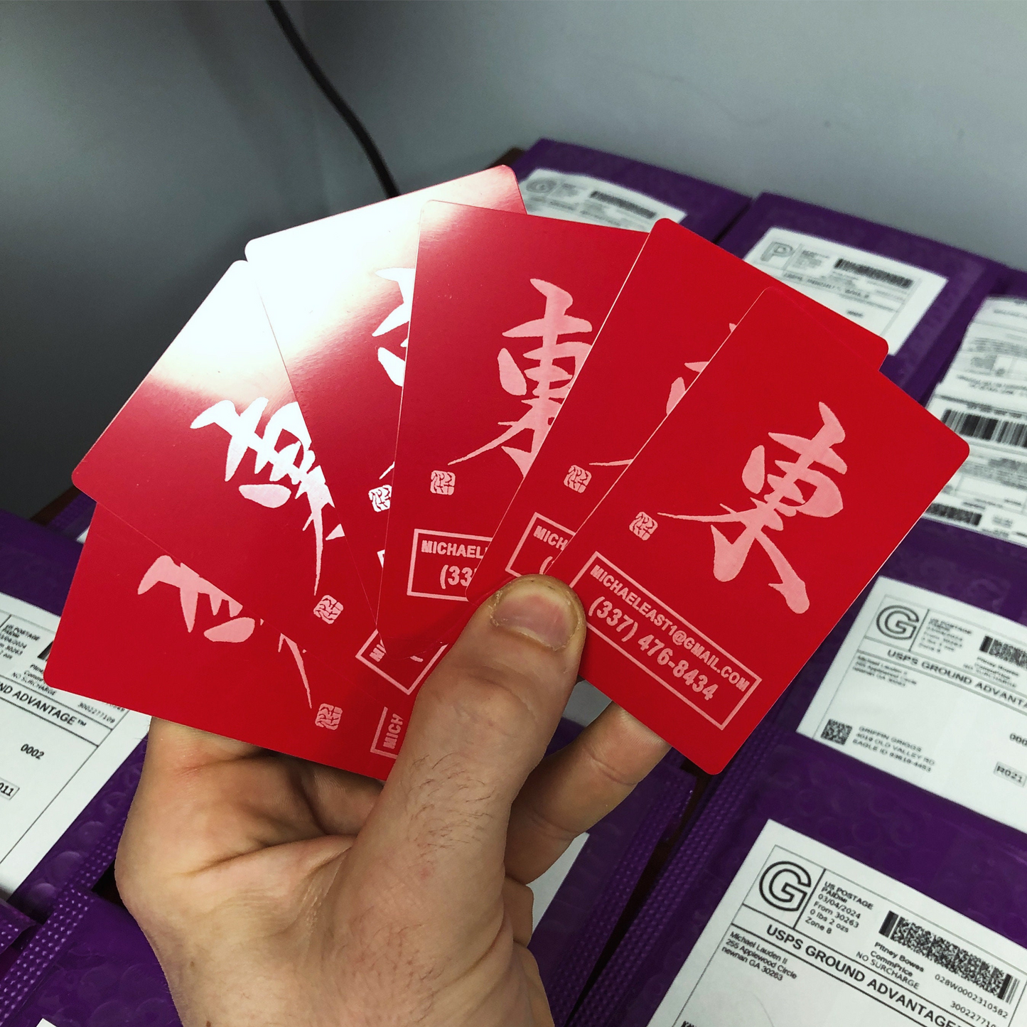 Hand holding a set of red/silver metal business cards with japanese kanji characters and customized contact information in front of rows of product mailers