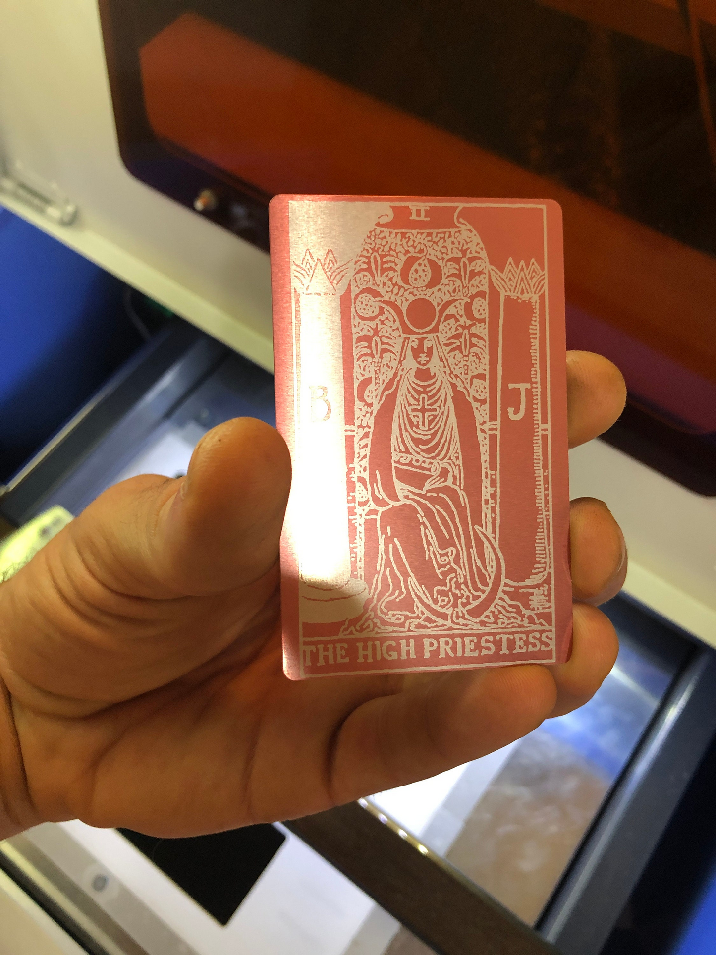 Hand holding a metal laser engraved tarot card with 'The High Priestess' design in front of a machine.
