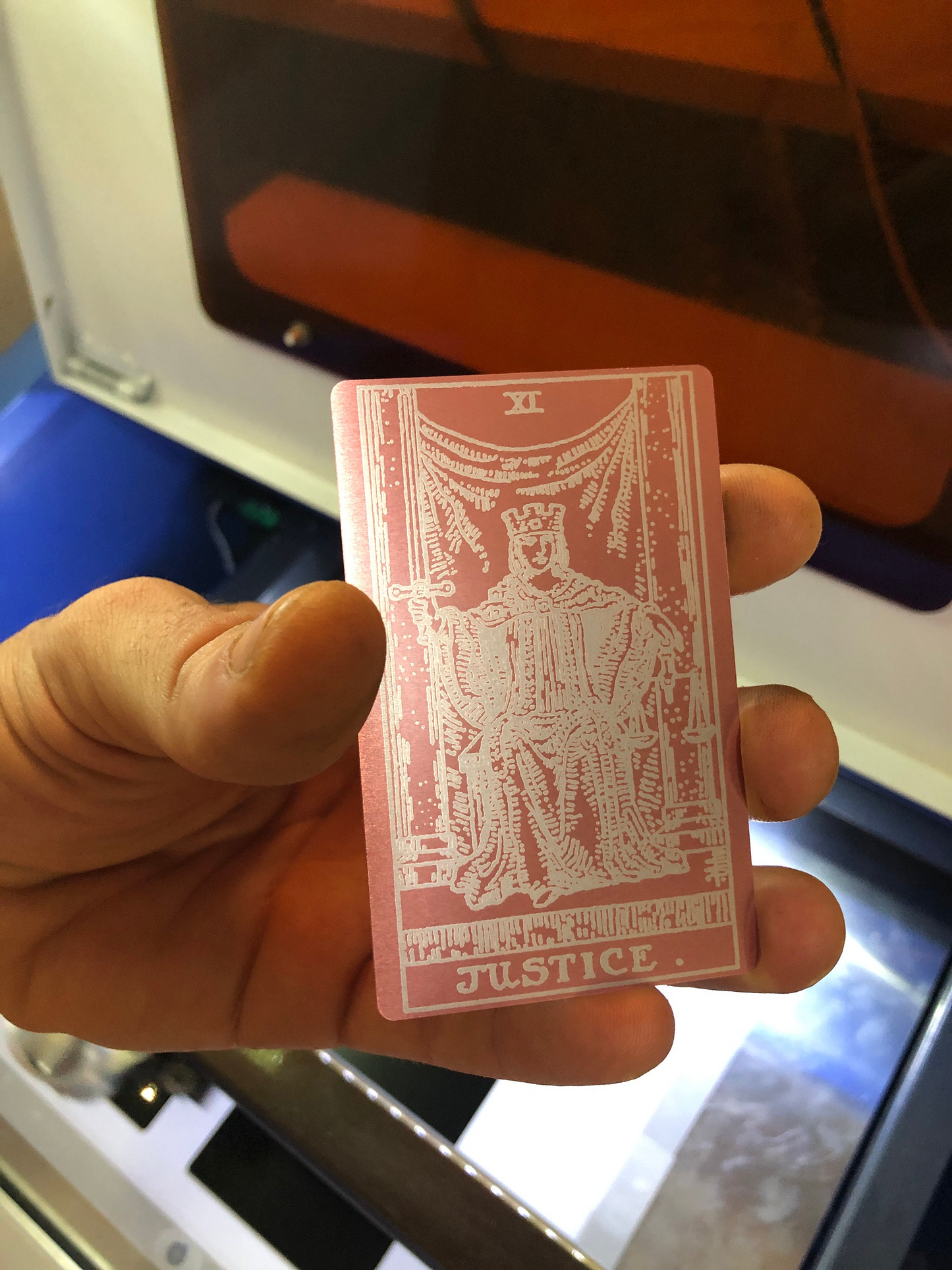 Hand holding a metal rose gold/silver tarot card with 'Justice' symbol against a blurred background