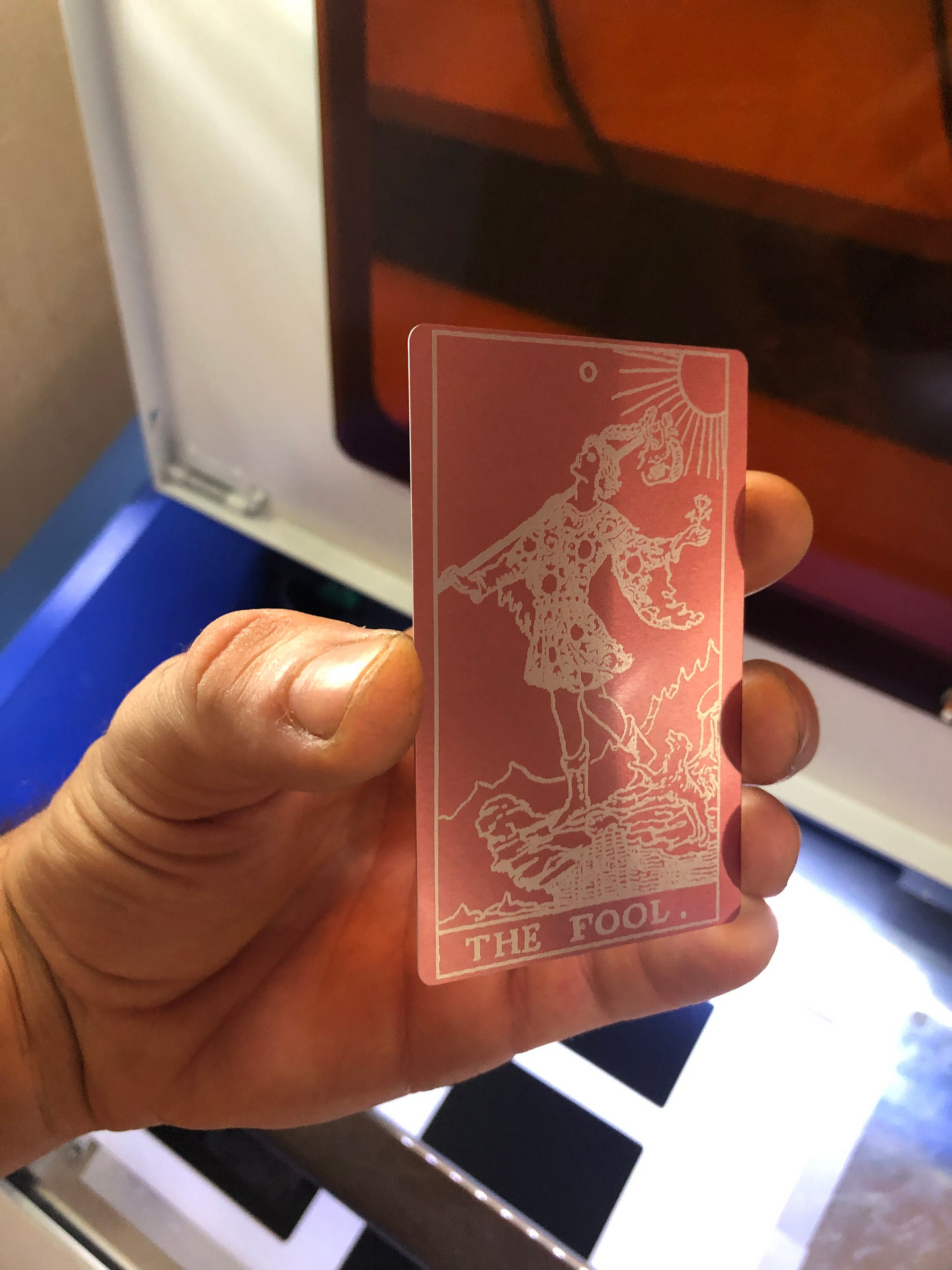 Hand holding a rose gold and silver metal keepsake card with a The Fool tarot card design labeled 'The Fool' against a blurred background.