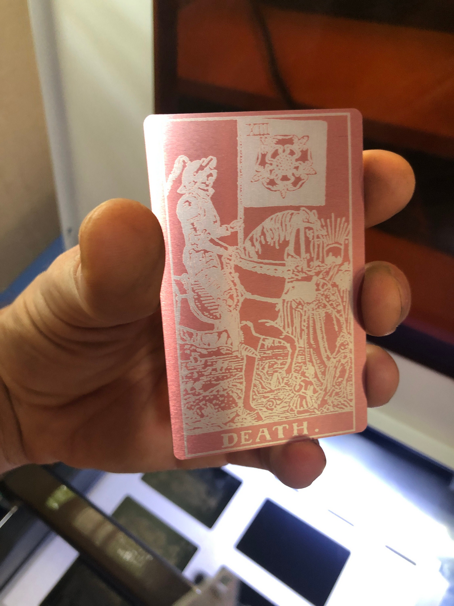 Hand holding a rose gold/silver metal tarot card with intricate designs and 'DEATH' text.