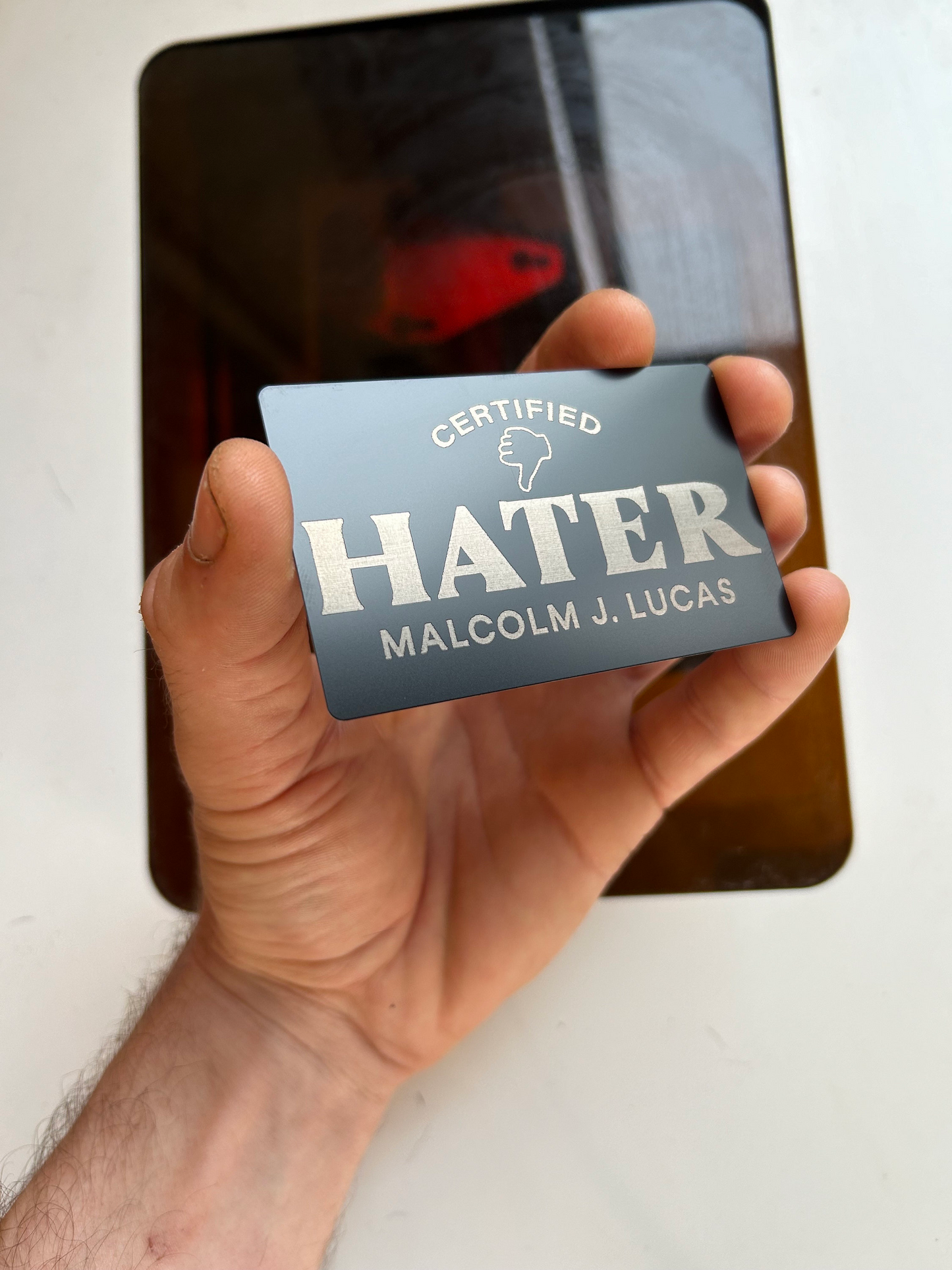Hand holding a card with 'Certified Hater' text against a blurred industrial machine background