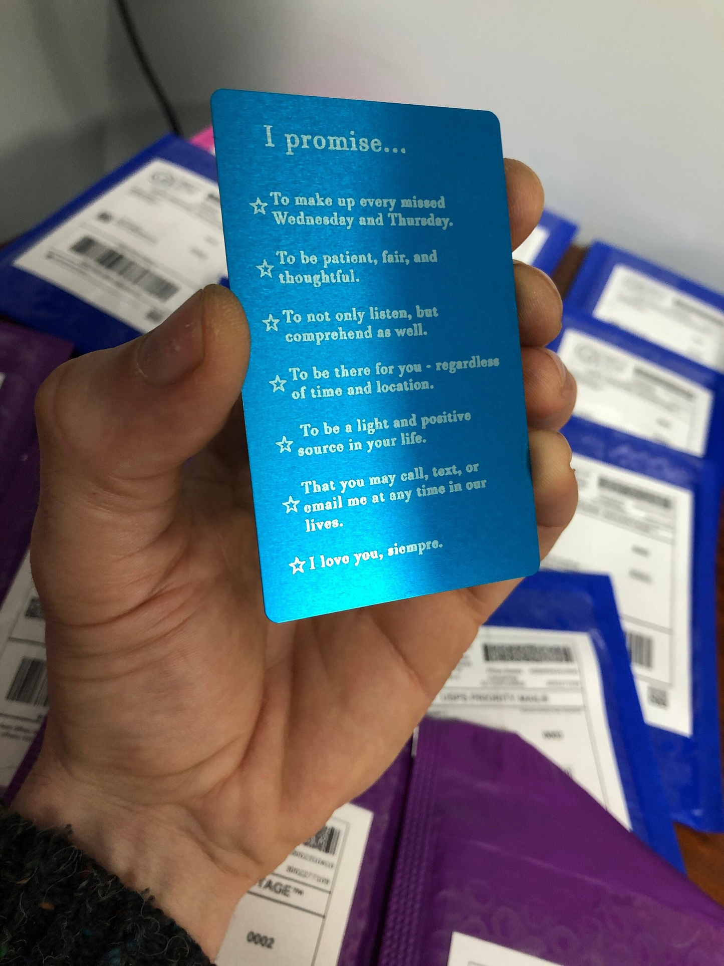 Hand holding a blue laser engraved anodized aluminum metal card with personalized "I Promise to...." text against a blurred background of product mailers