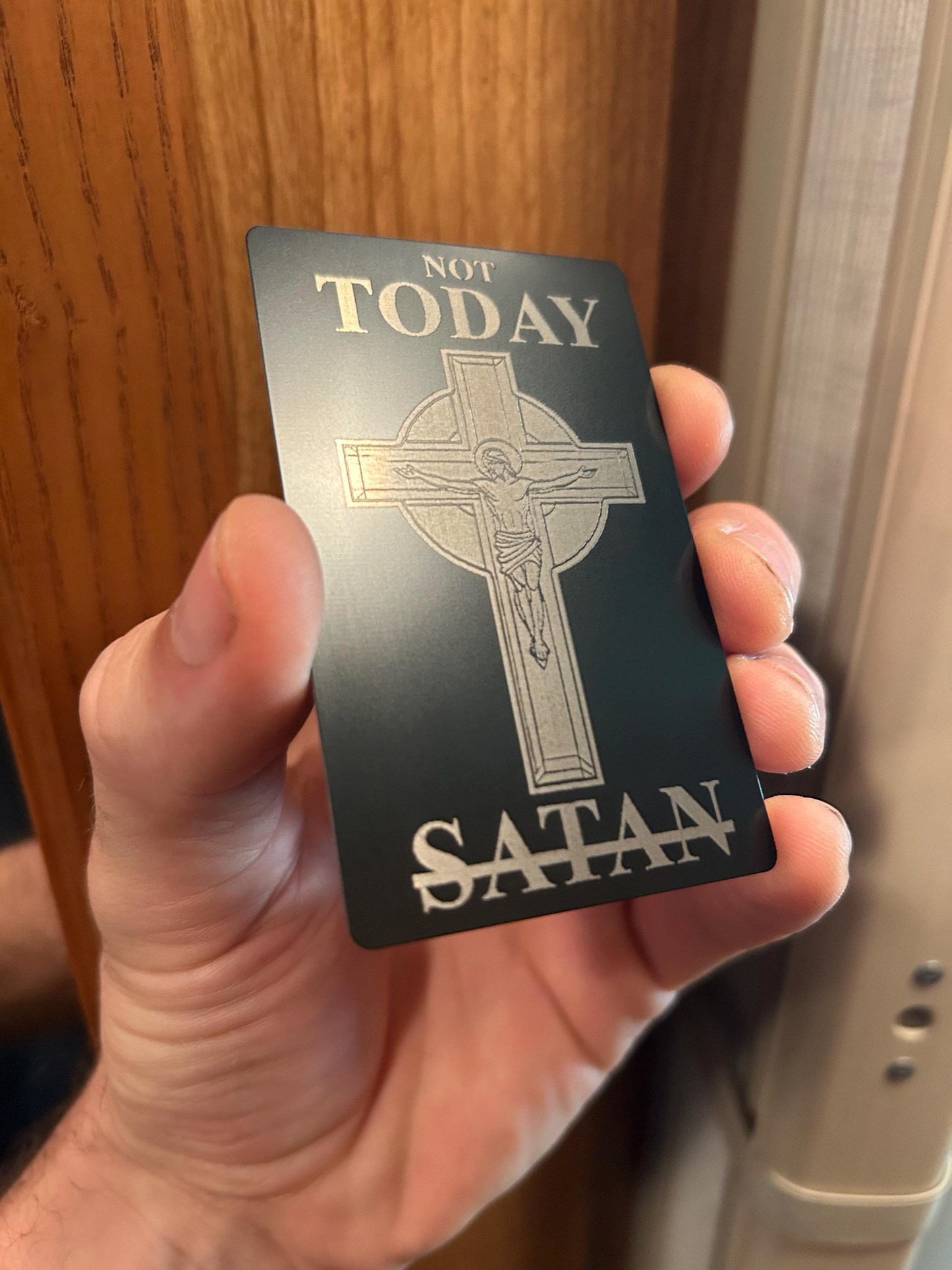 Hand holding a metal bookmark card with a cross design and text 'Not Today Satan' against a wooden door background.