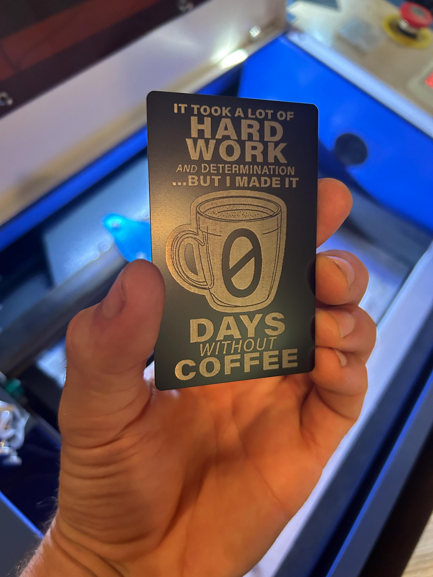 Hand holding a black/silver metal card with silver text and coffee graphic, set against a blurred machine background.