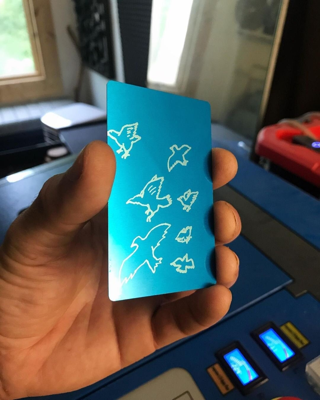 Hand holding a blue/silver metal anodized business card with hand drawn bird illustrations in front of a cnc machine