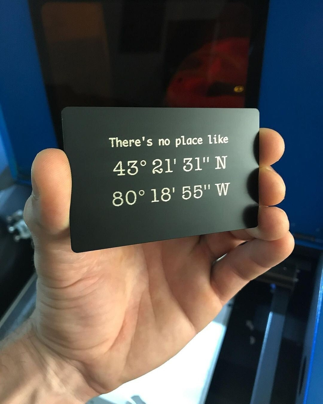 Hand holding an anodized aluminum keepsake placard with latitude longitude coordinates on a blurred background