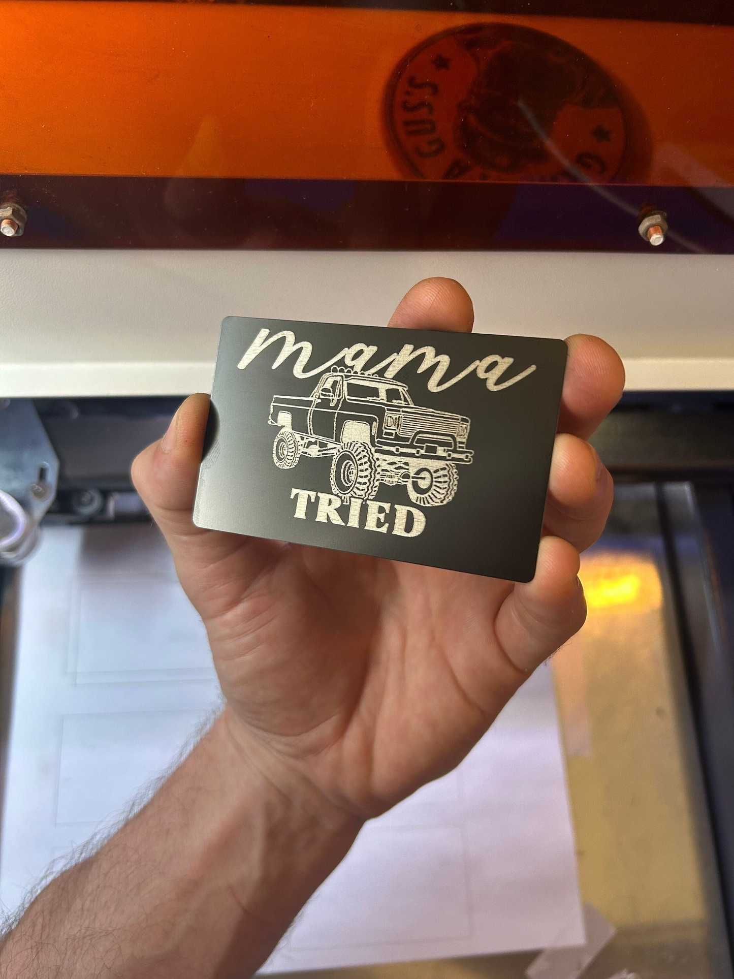 Hand holding a card with 'mama tried' text and truck illustration in front of a machine.