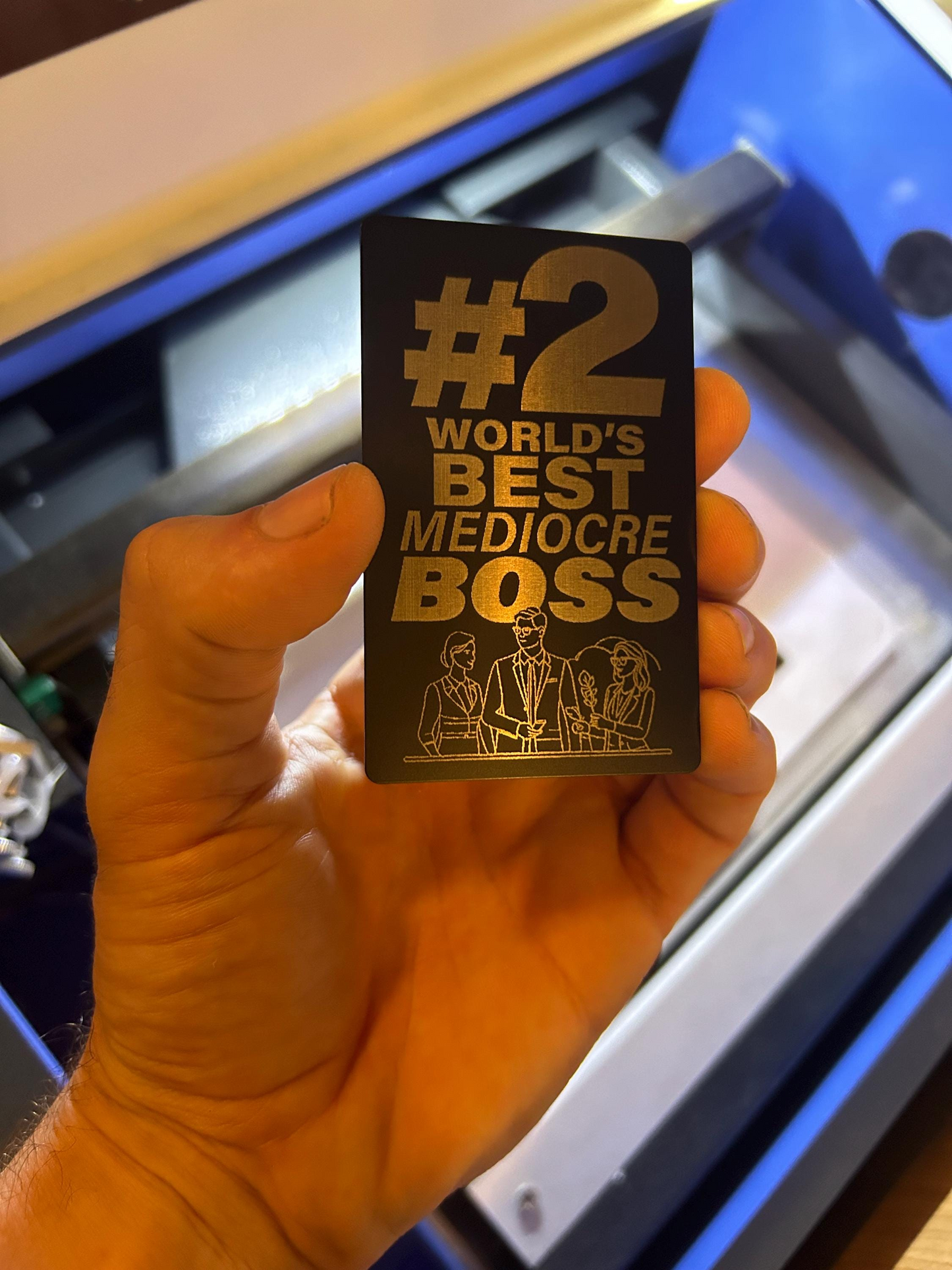 Hand holding metal keepsake #2 world's best mediocre boss card in front of a machine background