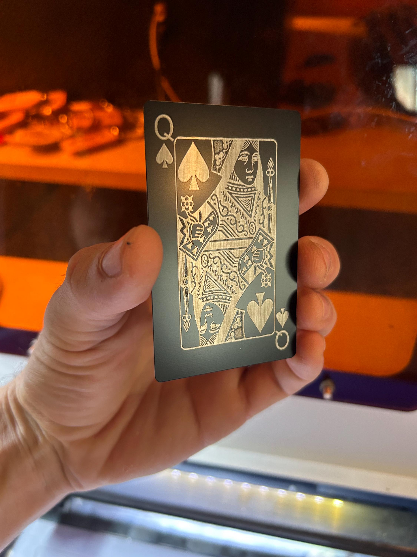 Hand holding a black/silver metal playing card with queen of spades design against a blurred background