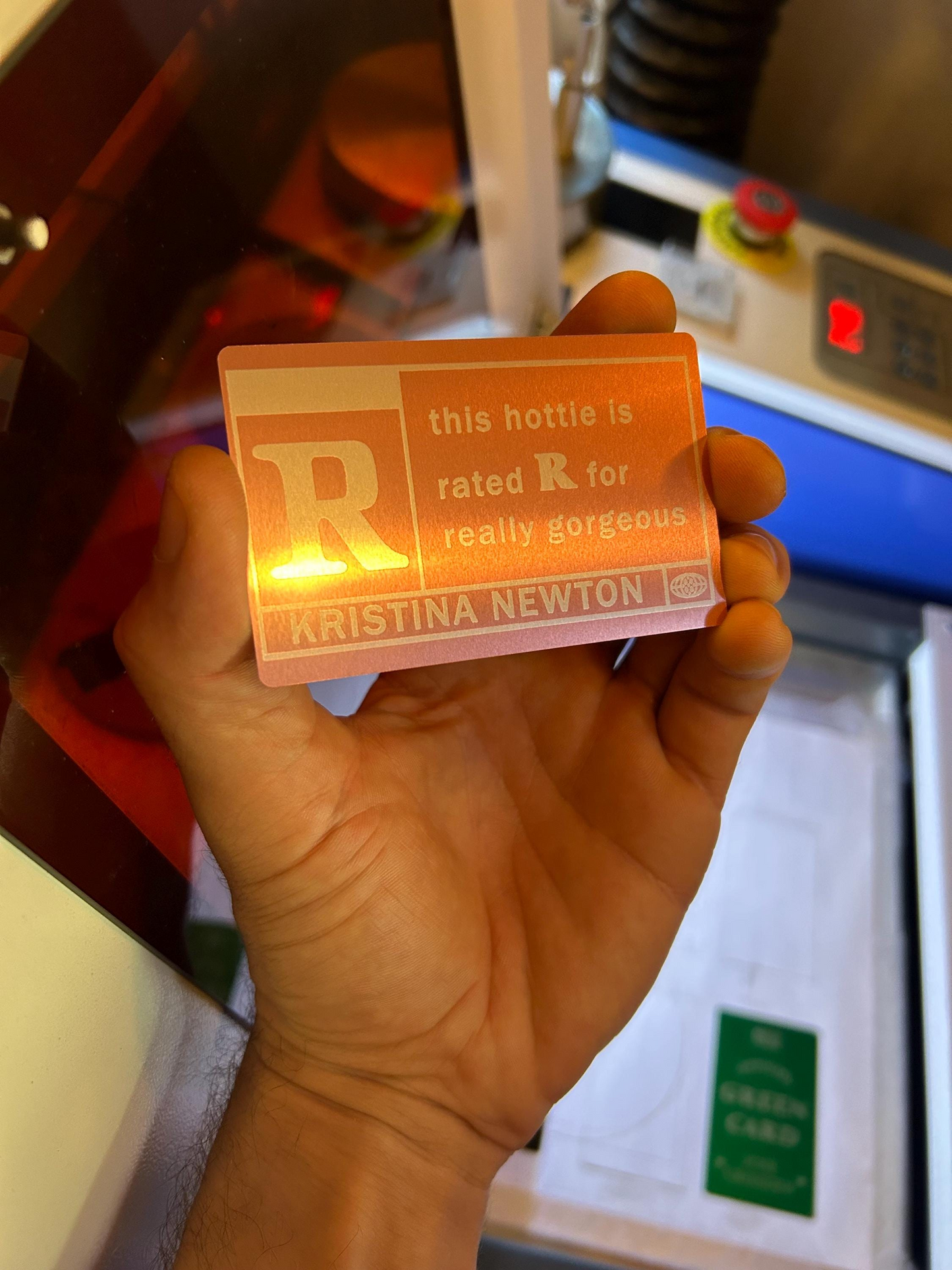 Hand holding a custom card engraved with custom text and a rating 'R' held in front of a laser engraving machine