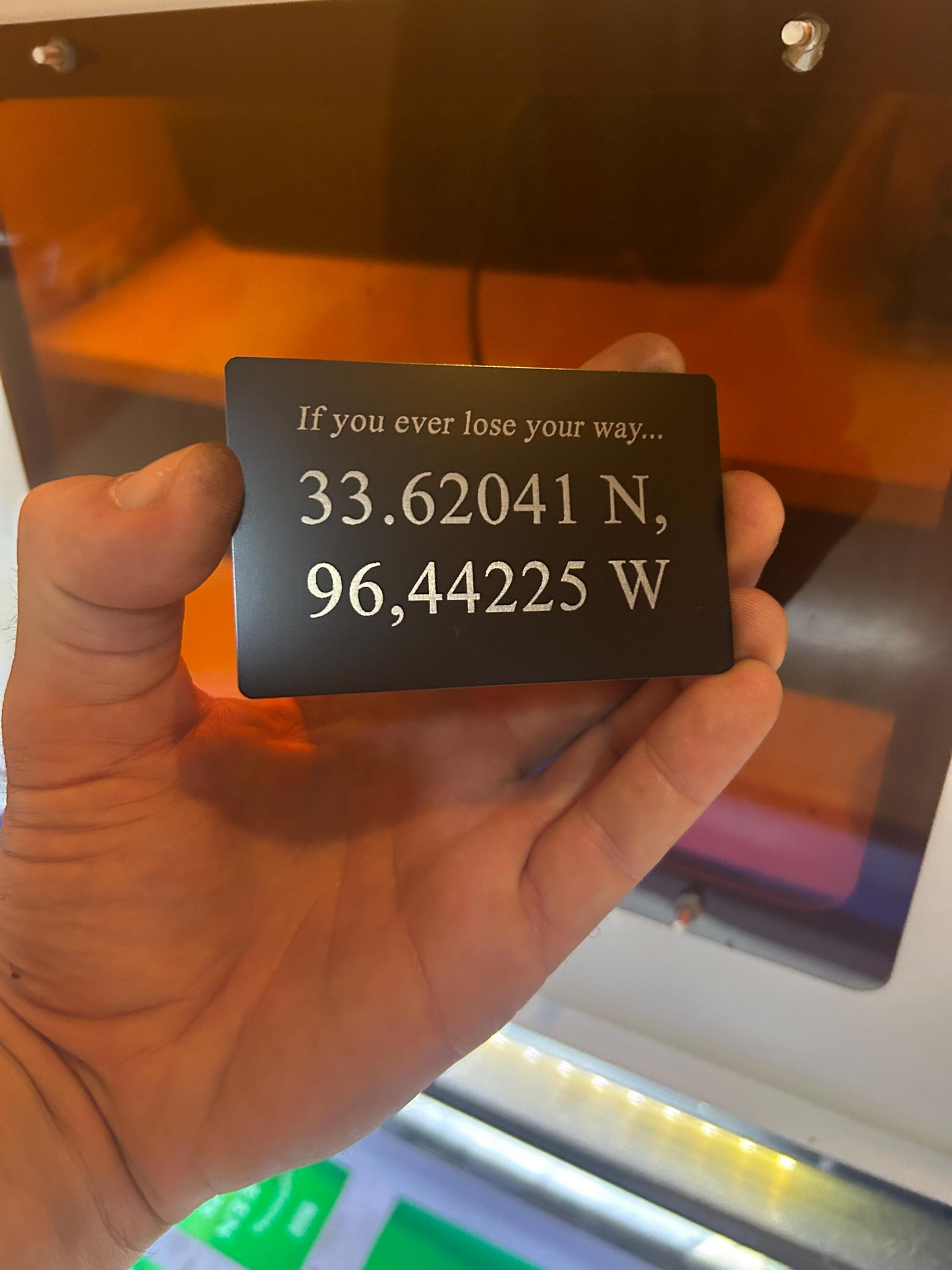 Hand holding a metal decorative plaque with GPS coordinates laser engraved, in front of a blurred background