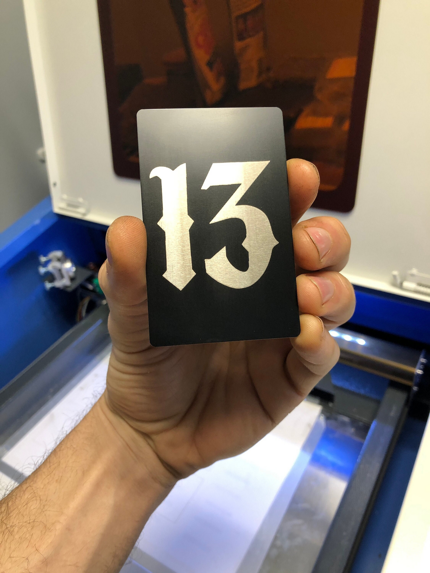 Hand holding a metal black/silver laser engraved luck charm card with the number 13 in front of a machine.