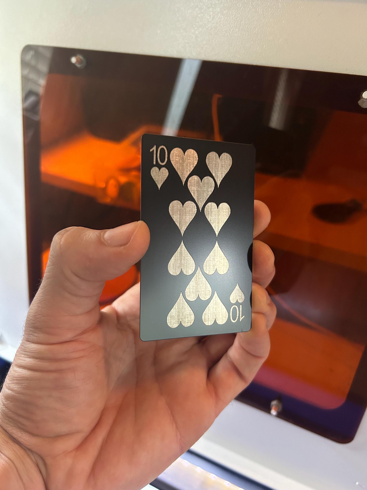 Hand holding an anodized aluminum laser engraved metal playing card with silver 10 of hearts design in front of a CNC laser engraving machine