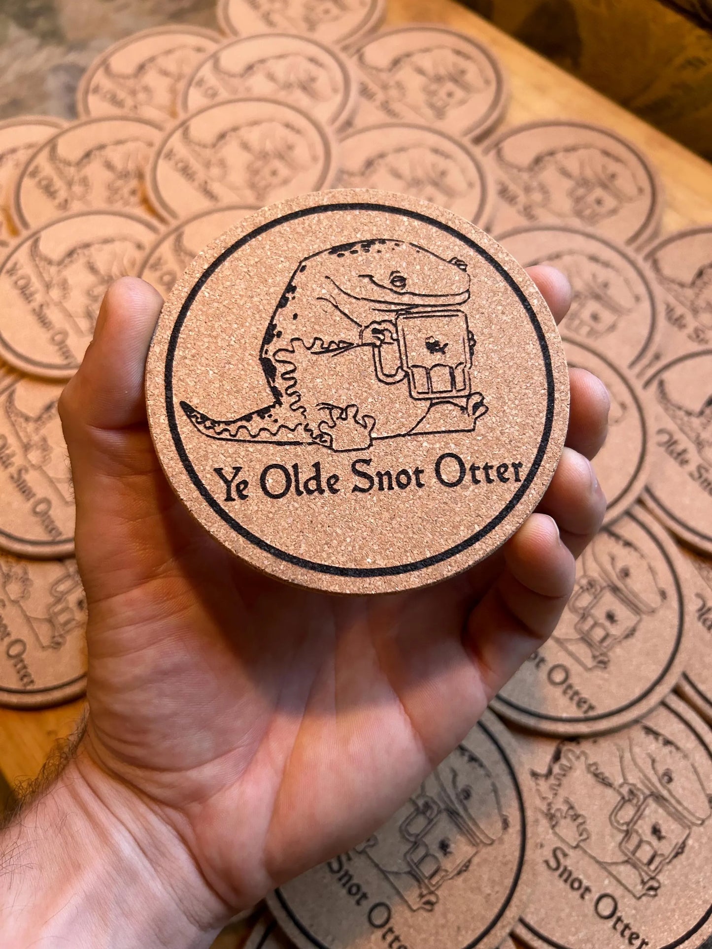 Coaster with 'Ye Olde Snot Otter' design held by a hand, surrounded by more coasters on a wooden surface.