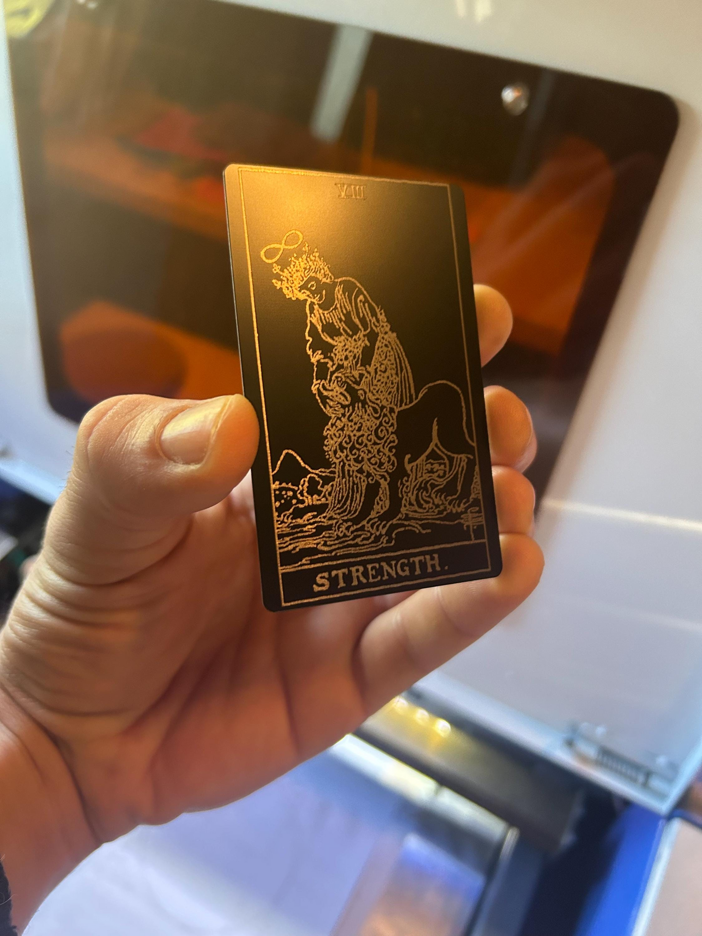 Hand holding a laser engraved metal tarot card with 'Strength' at the bottom, against a reflective surface.