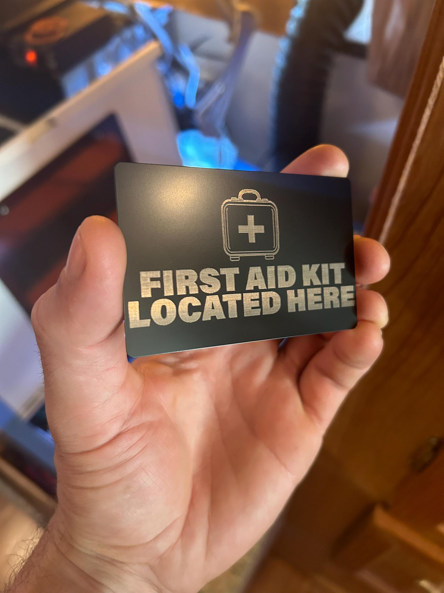 Hand holding a metal black placard with 'First Aid Kit Located Here' text and a medical symbol.