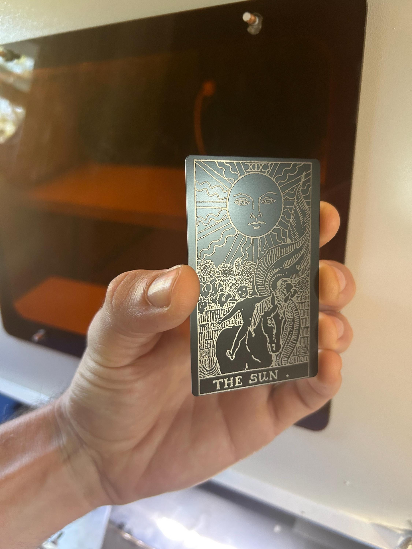Hand holding a metal tarot card titled 'The Sun' design laser engraved in front of an oven.