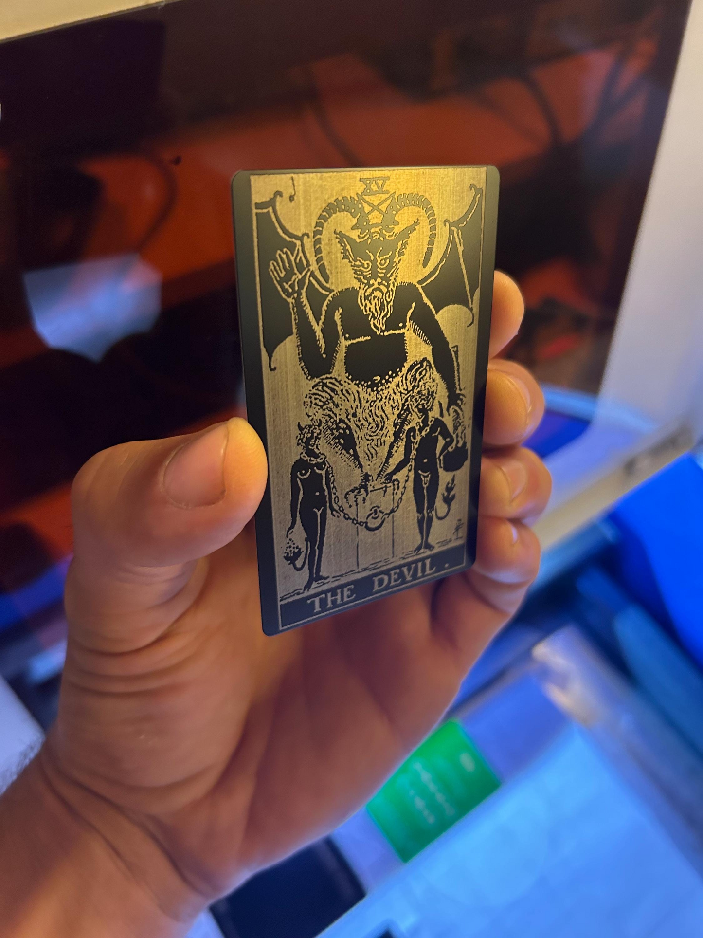 Hand holding a metal black/silver tarot card with 'The Devil' illustration against a blurred indoor background