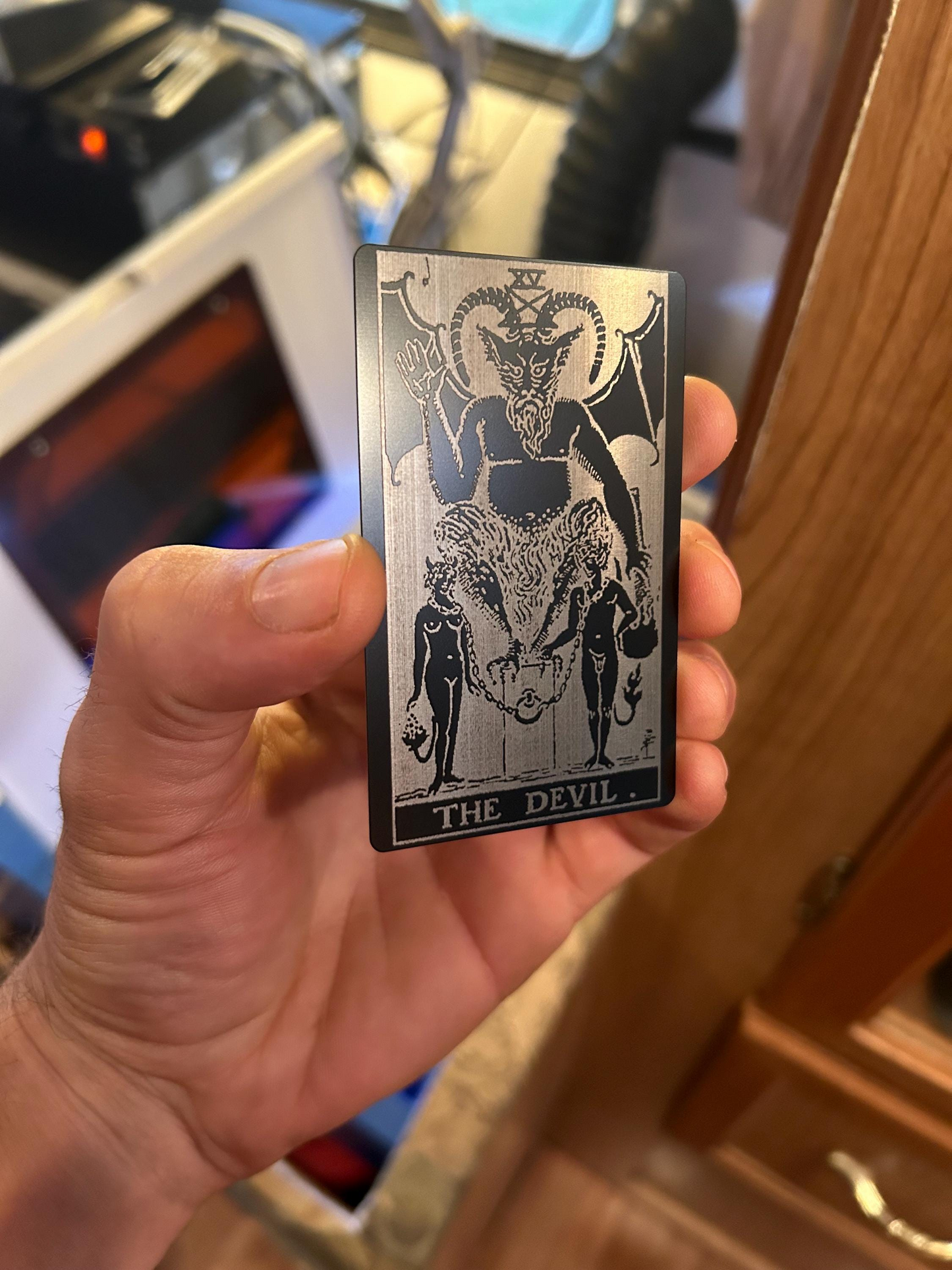 Hand holding a laser engraved metal tarot card titled 'The Devil' in an indoor setting.