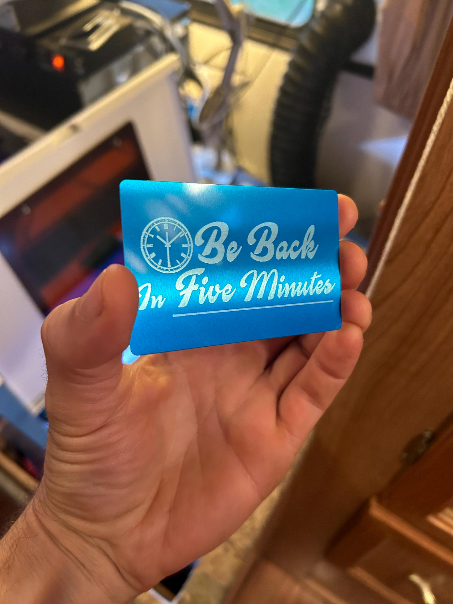 Hand holding a blue/silver metal card with 'Be Back In Five Minutes' text in an industrial setting