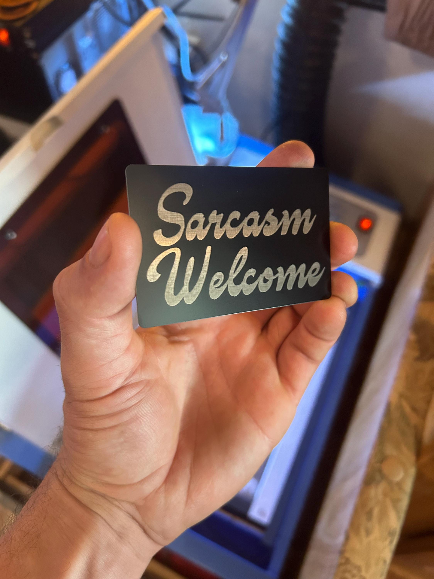 Hand holding a metal mini signage card with 'Sarcasm Welcome' text in front of a blurred background