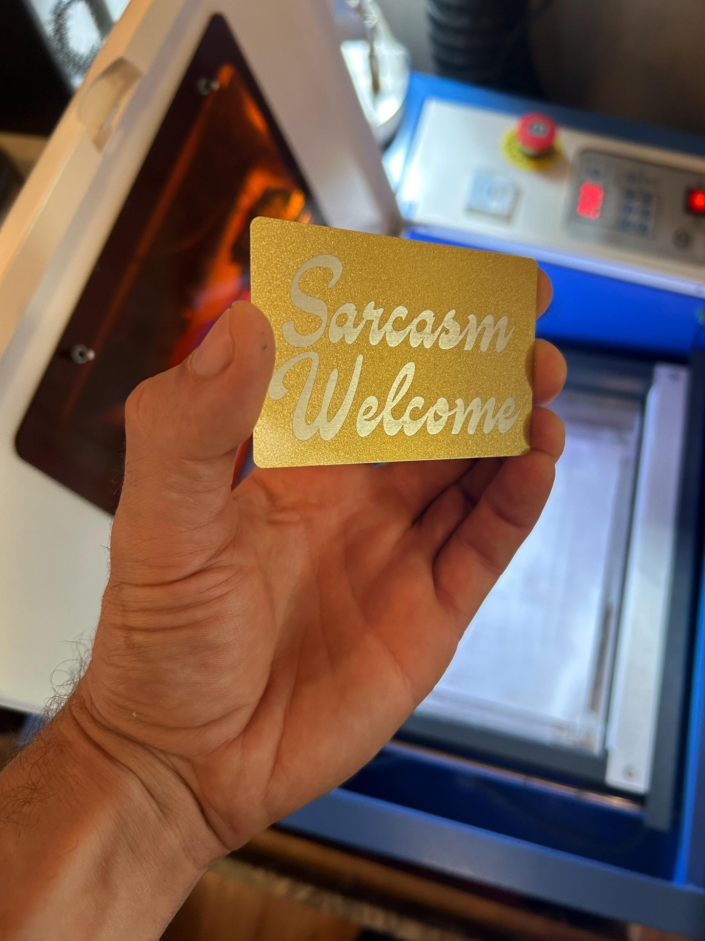 Hand holding a gold/silver metal card with 'Sarcasm Welcome' text in front of a machine.