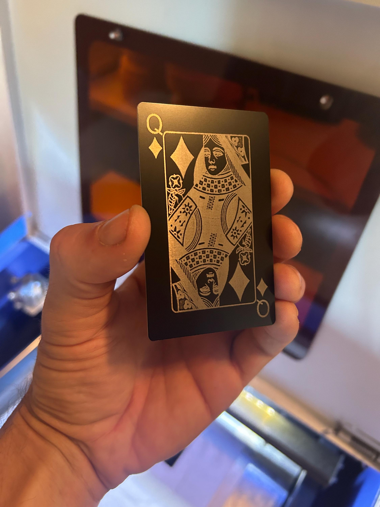 Hand holding a black metal queen of diamonds playing card with silver design against a machine background