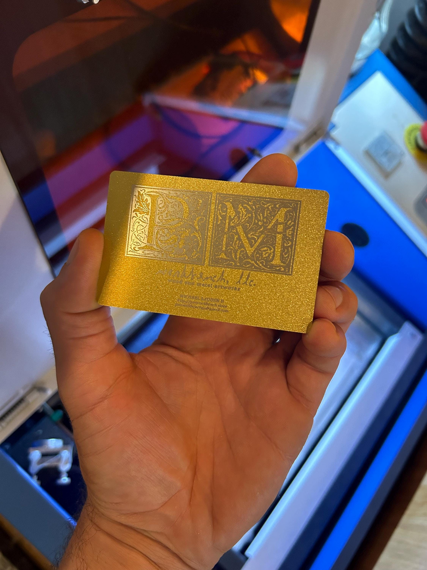 Hand holding a gold/silver metal business card with intricate design and contact information in front of a colorful background