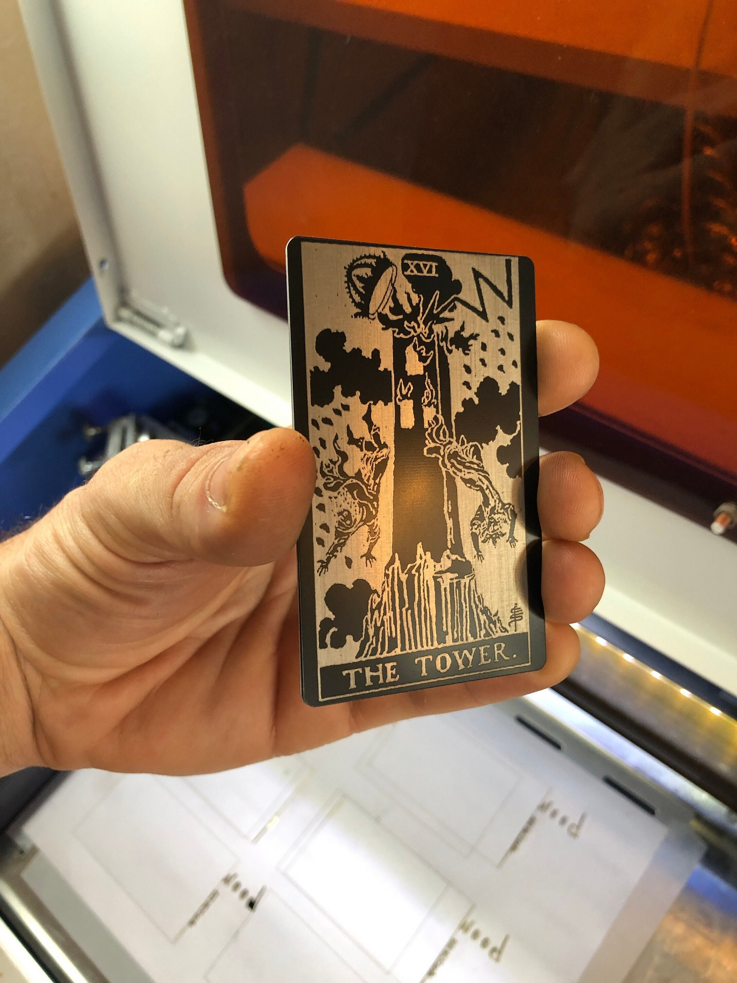 Hand holding a metal keepsake gift -- it is a laser engraved tarot card titled 'The Tower' in front of a machine.