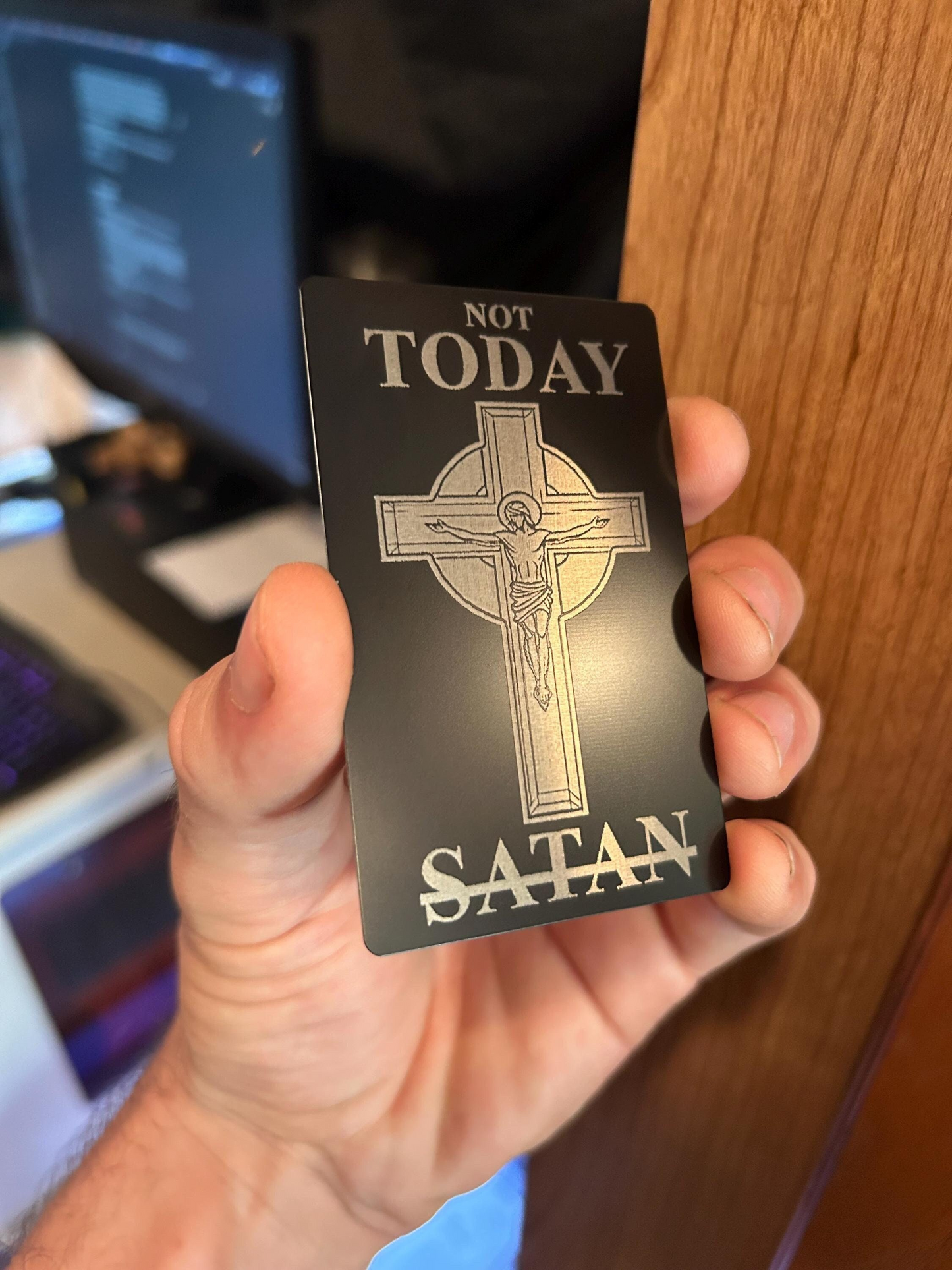 Hand holding a metal engraved card with a cross design and text 'Not Today Satan' in front of a blurred background.