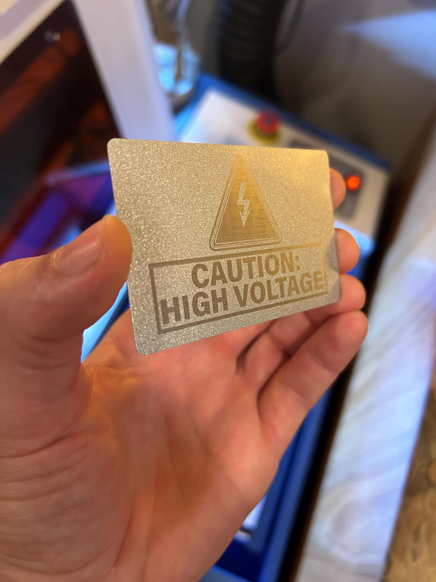 Hand holding a laser engraved silver/silver metal placard with a 'Caution: High Voltage' symbol