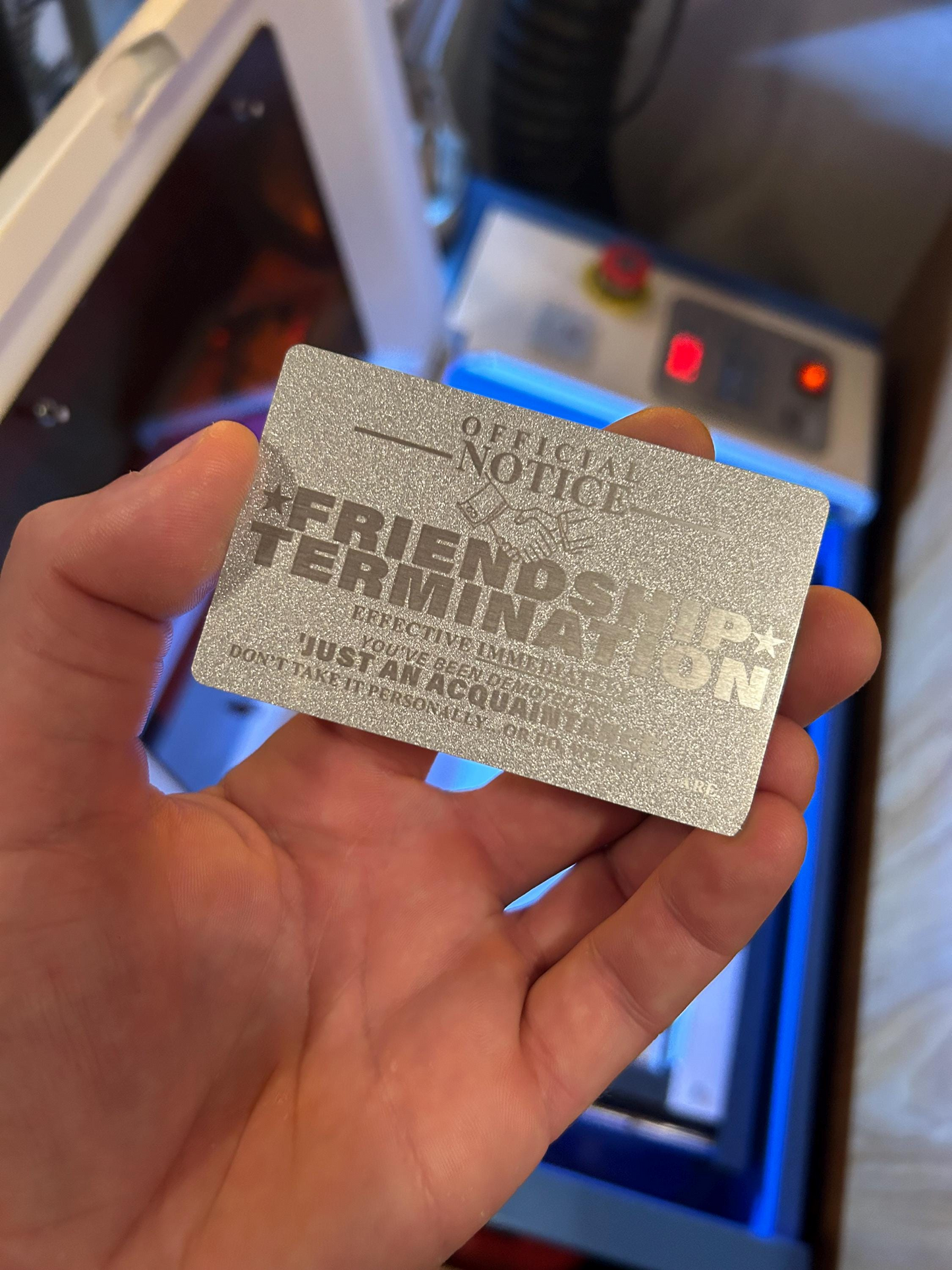 Hand holding a silver/silver laser engraved metal novelty gift card with text 'OFFICIAL NOTICE FRIENDSHIP TERMINATION' in front of a machine.