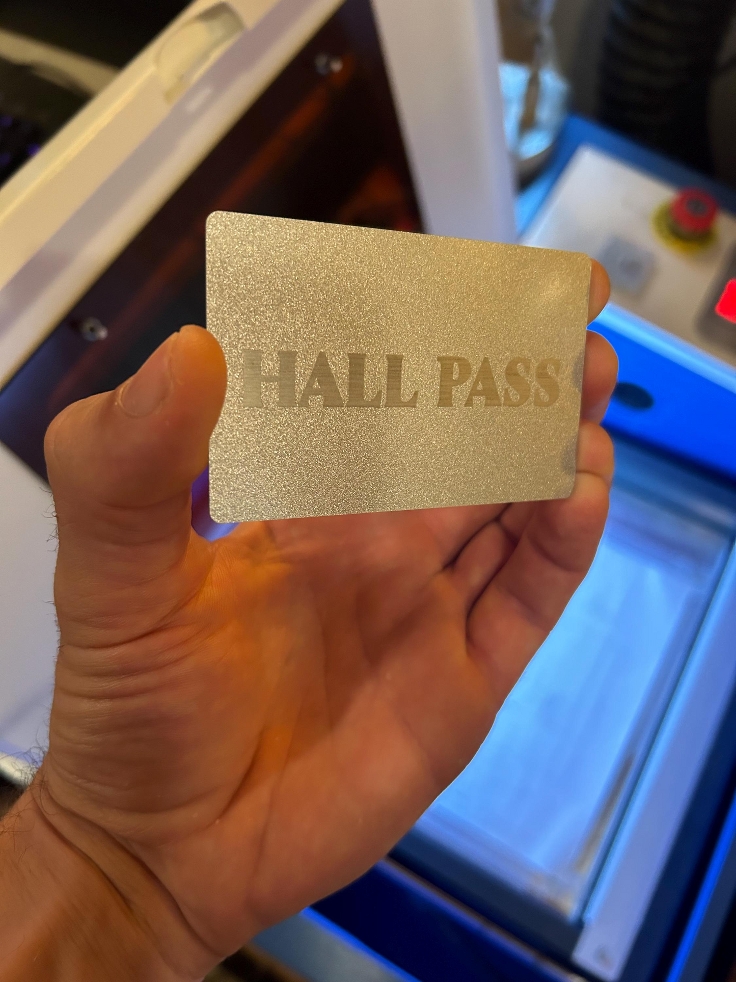 Hand holding a pearl silver 'HALL PASS' card in front of a machine.