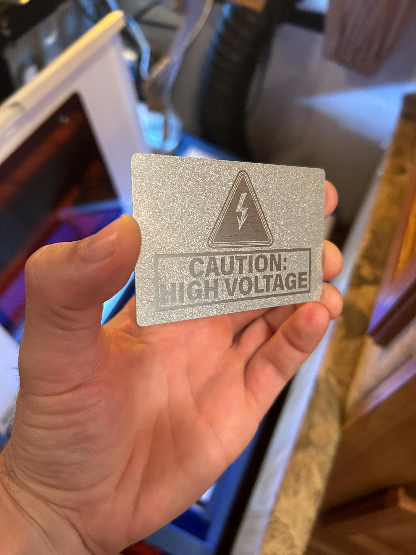 Hand holding a pearl silver laser engraved warning sign with a high voltage warning symbol
