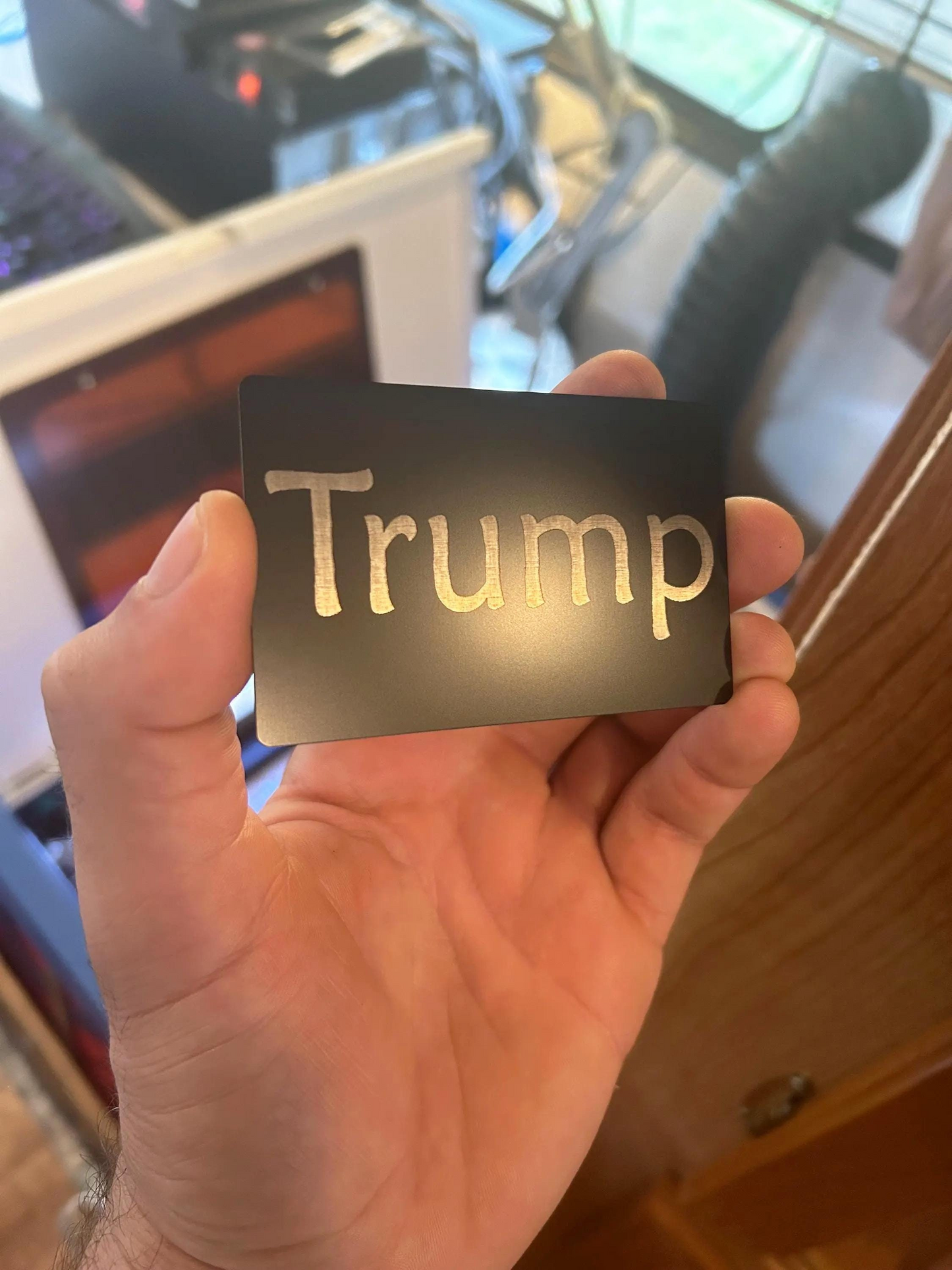 Hand holding a metal inside joke gift card with 'Trump' engraved on it in a casual indoor setting. "Trump Card" humor