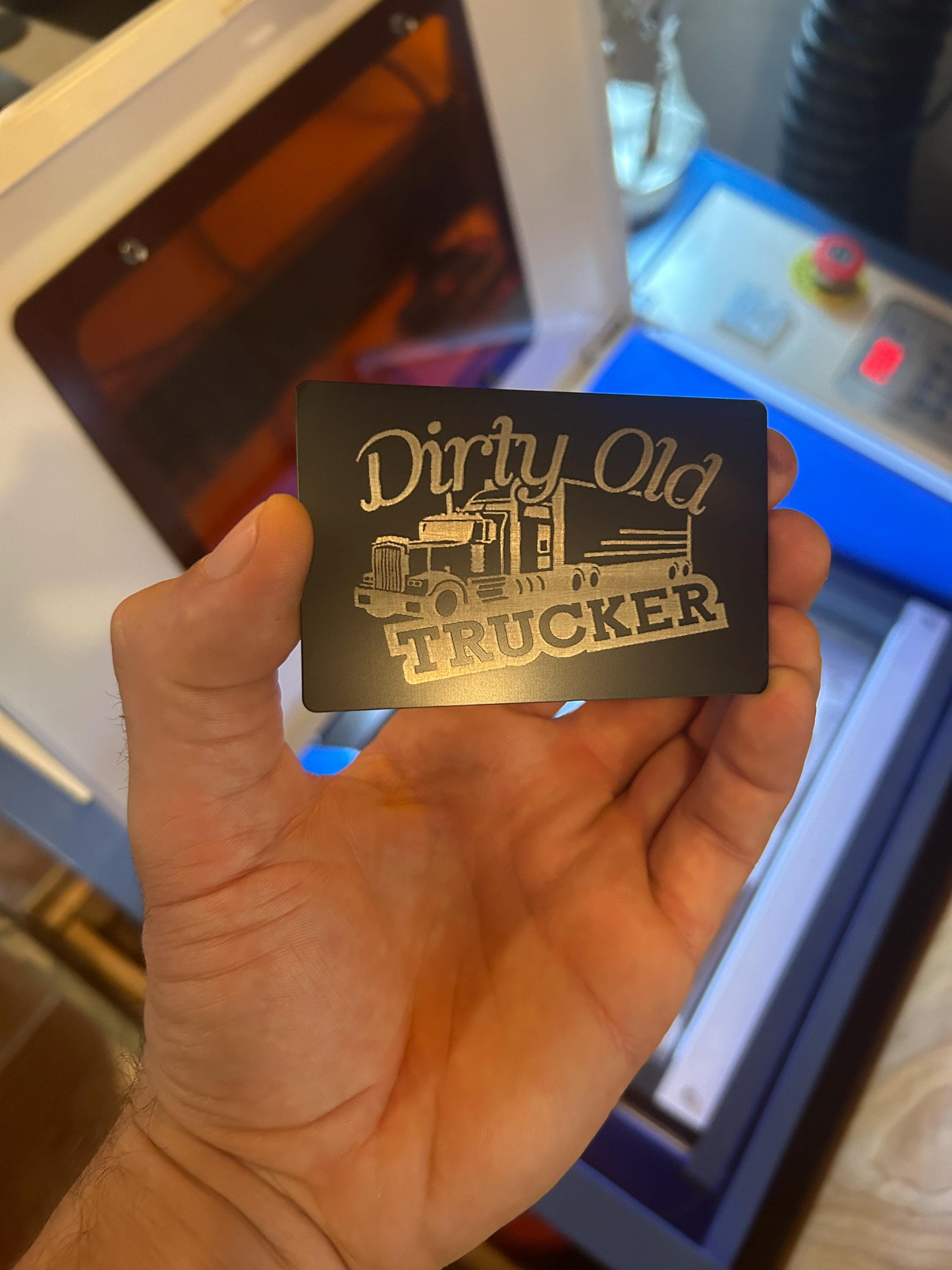 Hand holding a 'Dirty Old Trucker' card with a truck graphic in front of a machine.