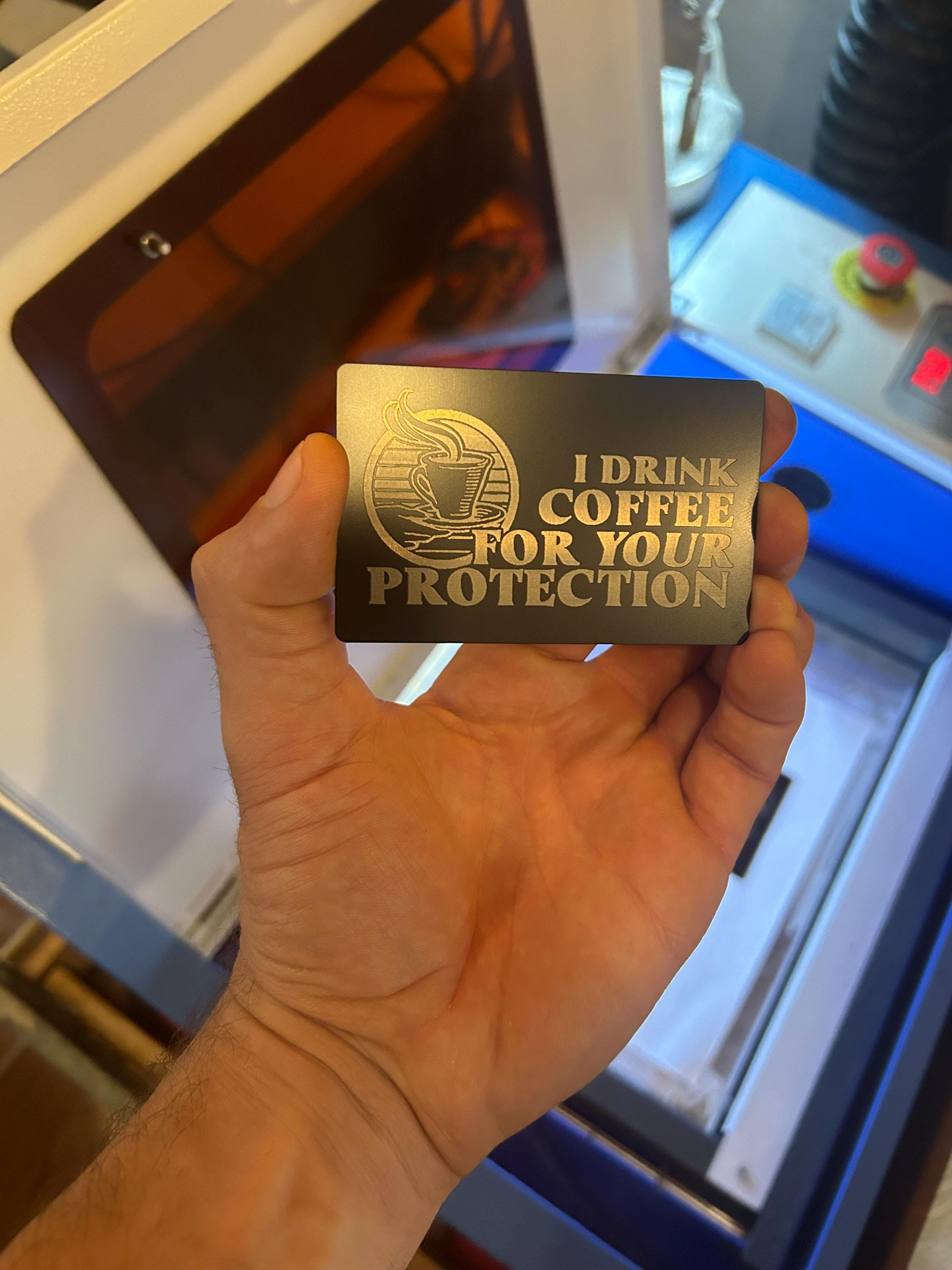 Hand holding a gold card with 'I Drink Coffee for Your Protection' text in front of a machine.