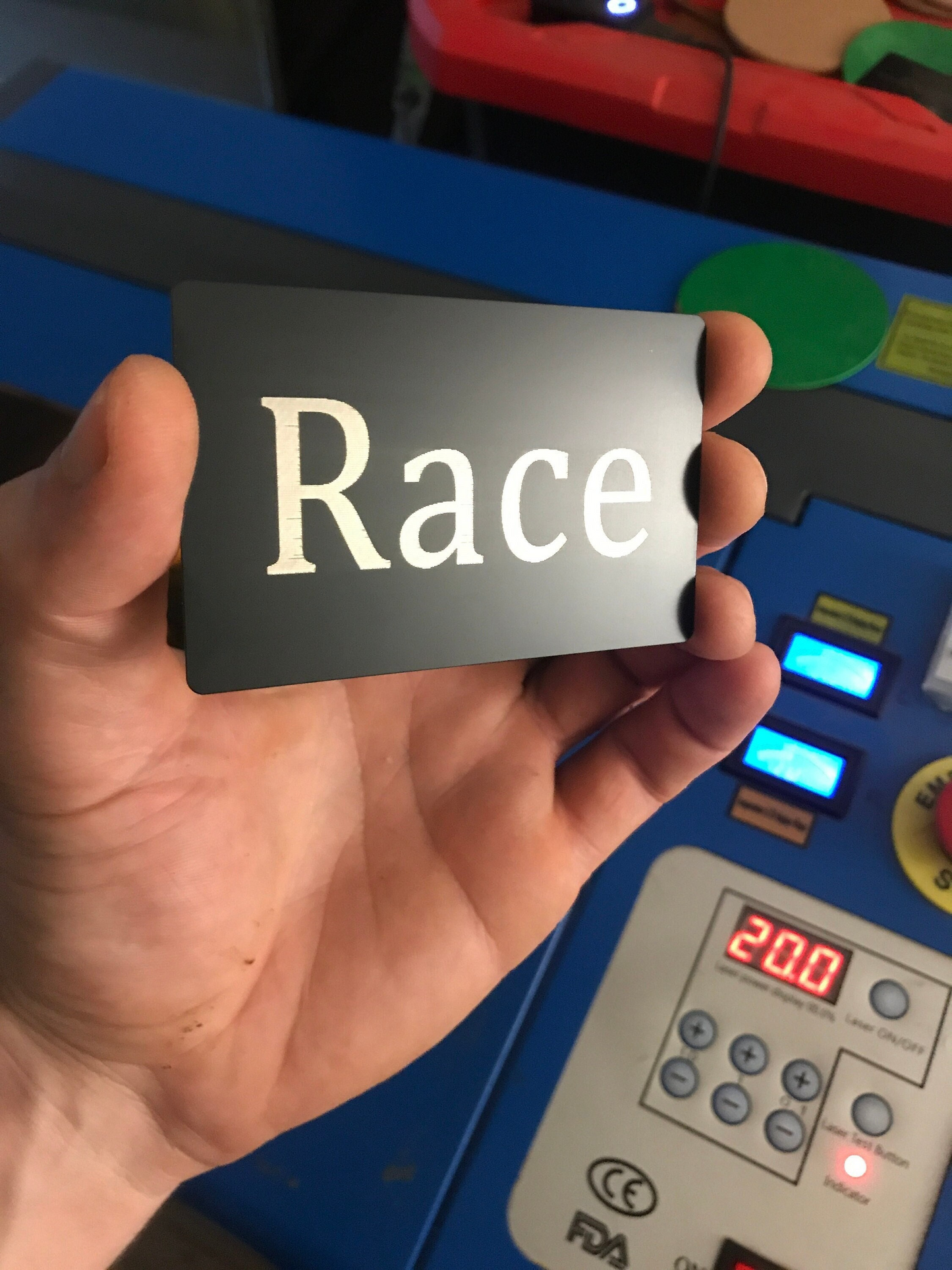 Hand holding a card with 'Race' laser engraved on it in front of electronic equipment.