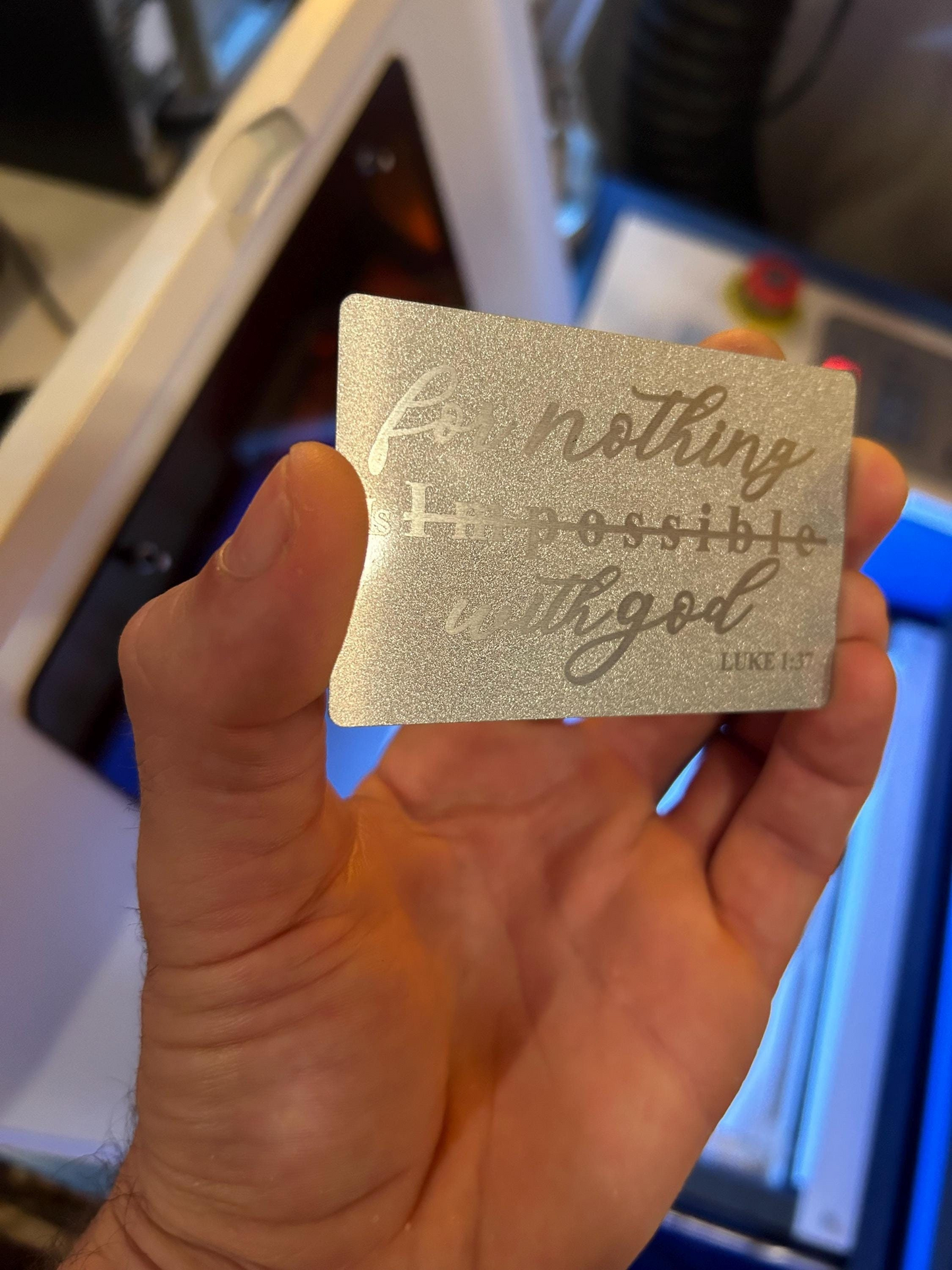 Hand holding a laser engraved silver on silver metal business card, with custom typography design in front of a cnc laser engraving machine