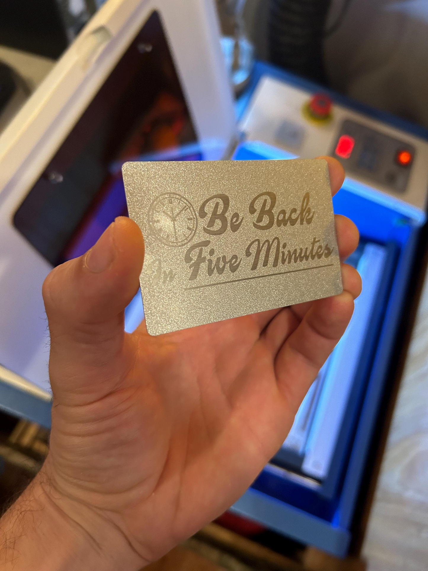 Hand holding a silver metal card with 'Be Back in Five Minutes' text in front of a machine.