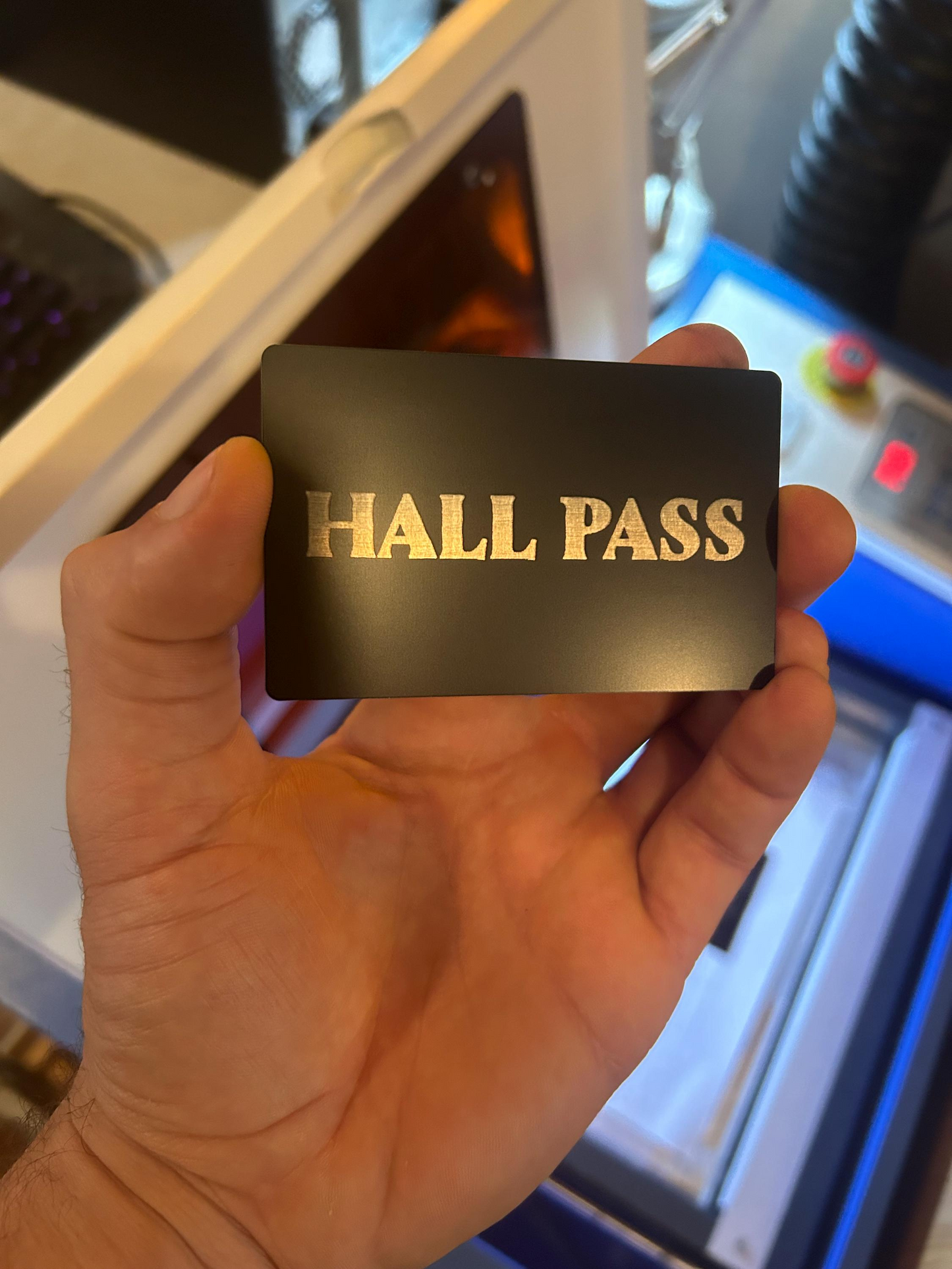 Hand holding a metal black card with 'HALL PASS' in silver letters against a blurred indoor background.