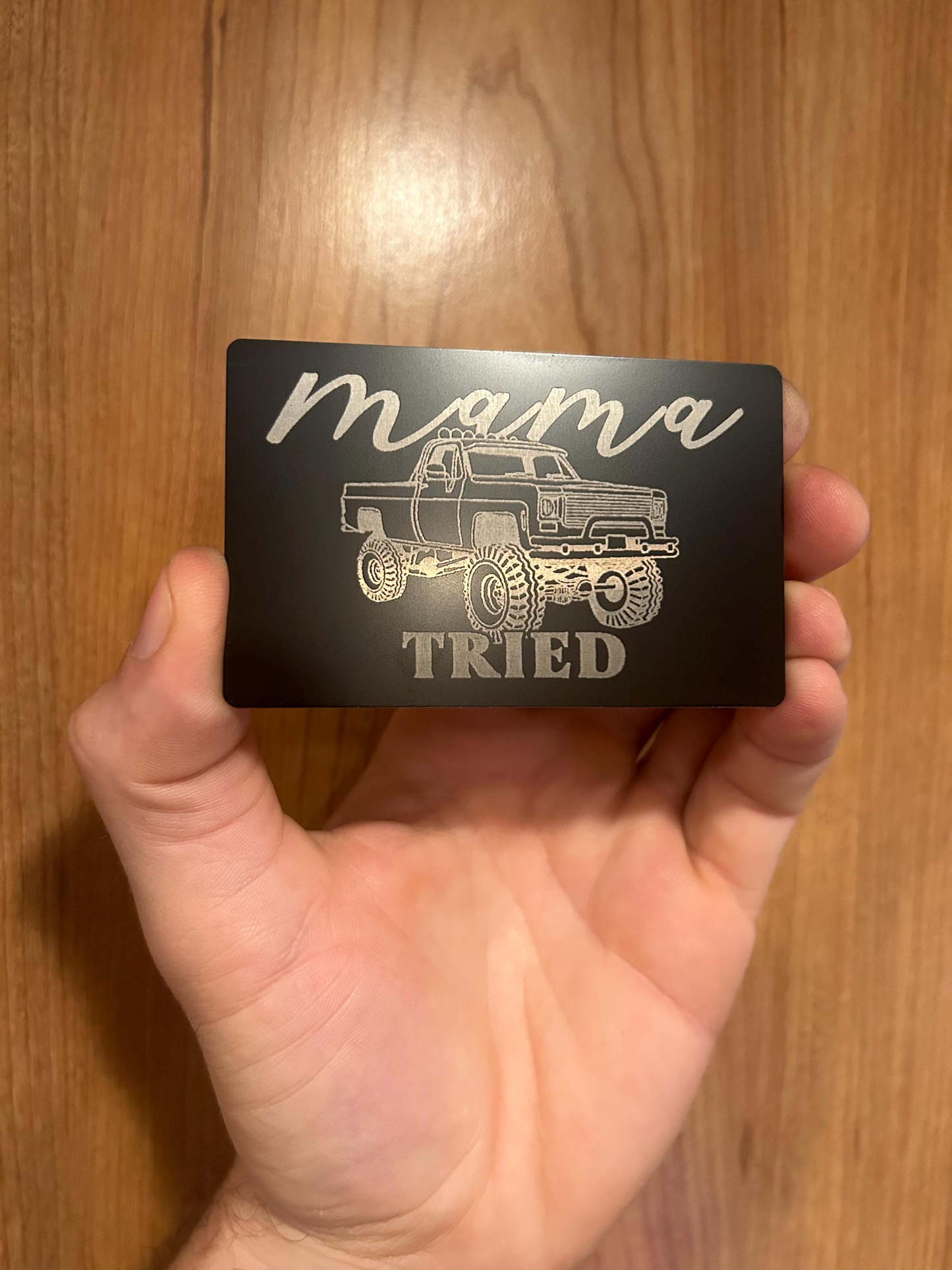 Hand holding a black card with 'mama tried' text and mudding truck illustration on a wooden surface