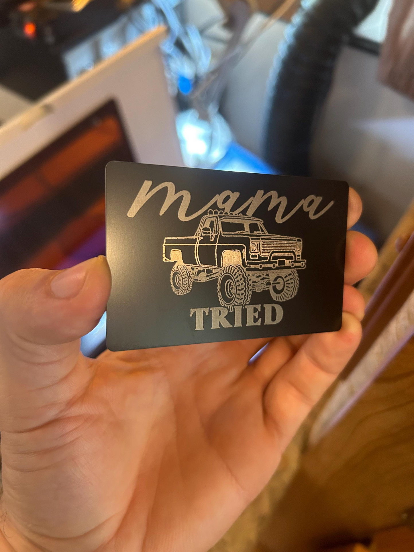 Hand holding a matte black and silver laser engraving with 'mama tried' and a truck illustration.