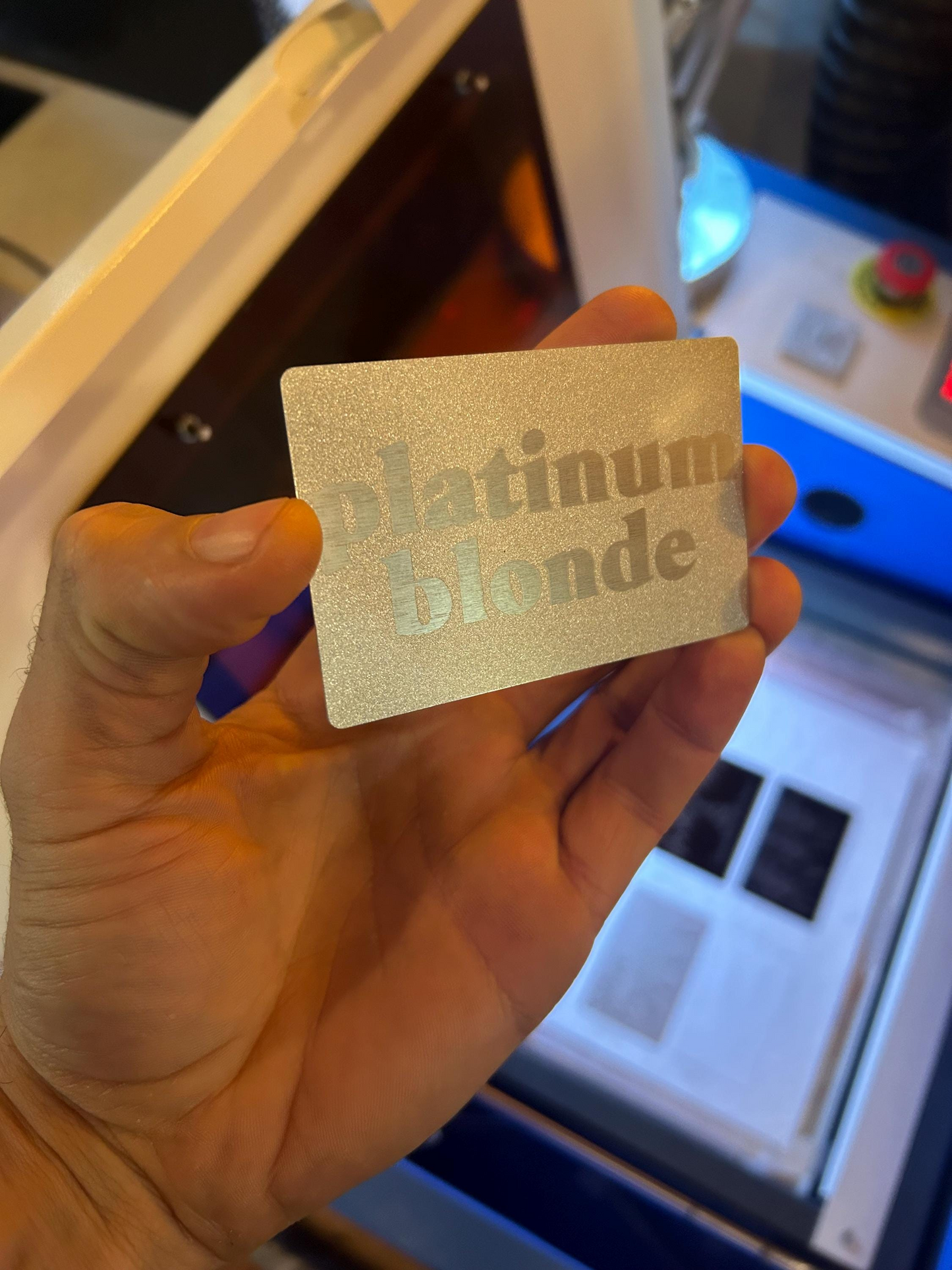 Hand holding a silver laser engraved funny keepsake card with 'platinum blonde' text in front of a blurred background